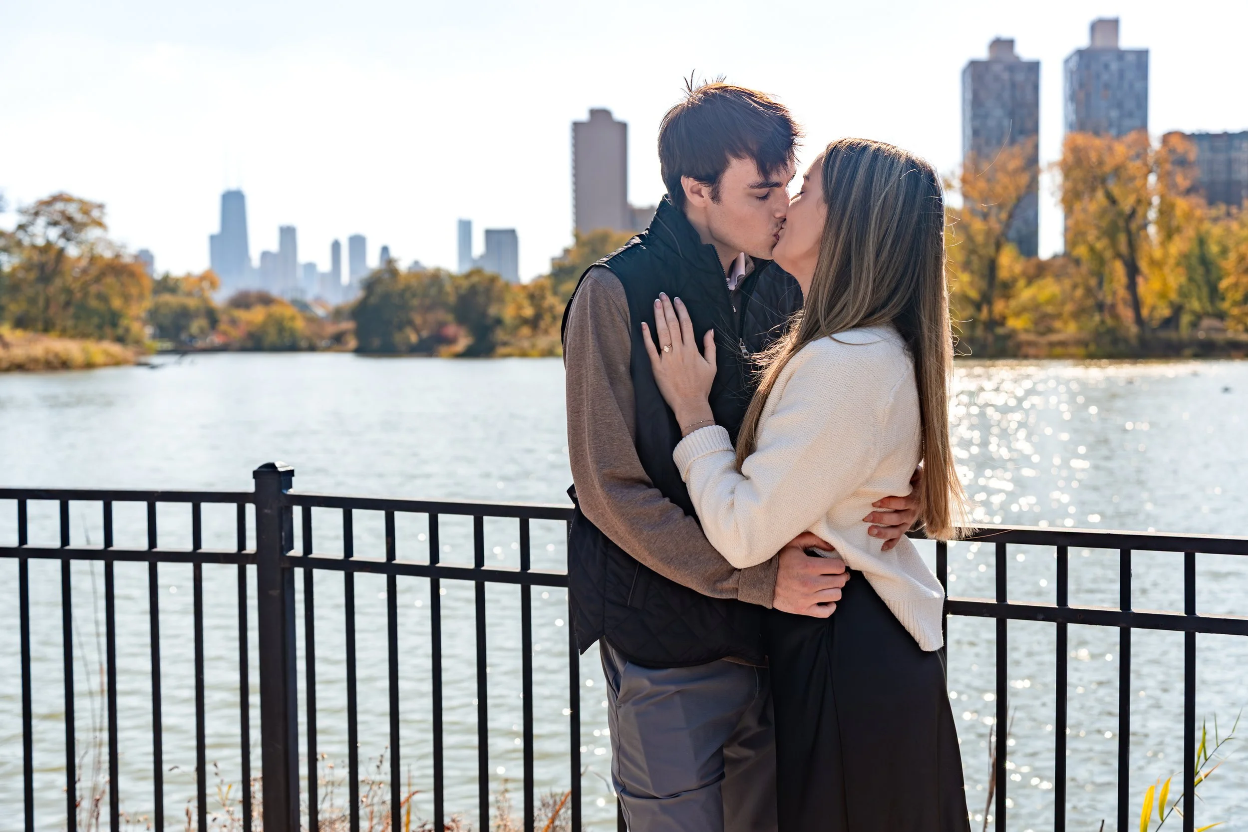 A couple kissing by a waterfront with city skyline and autumn trees in the background.