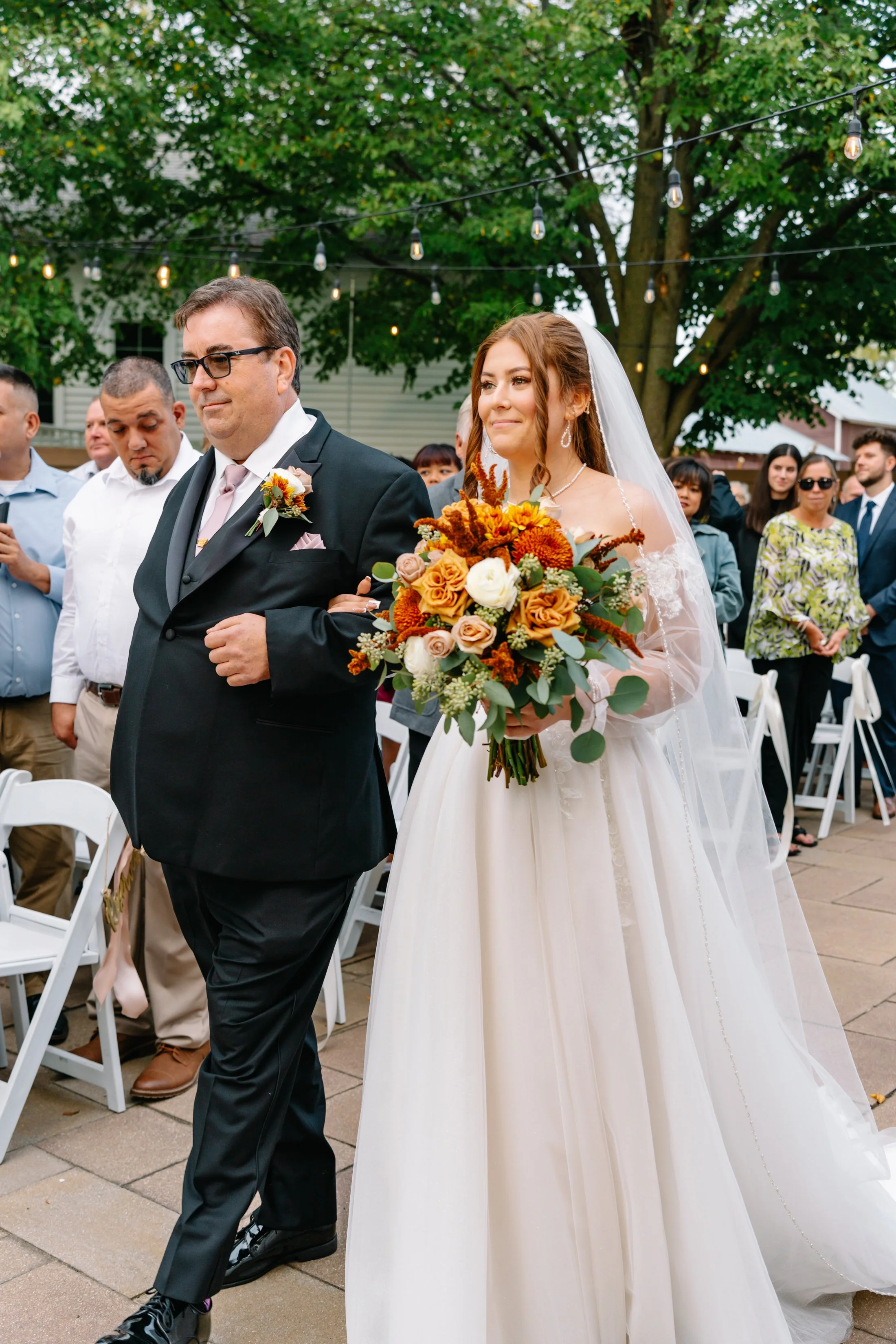 A bride walking down the aisle with her father at an outdoor wedding ceremony, holding a bouquet of flowers.