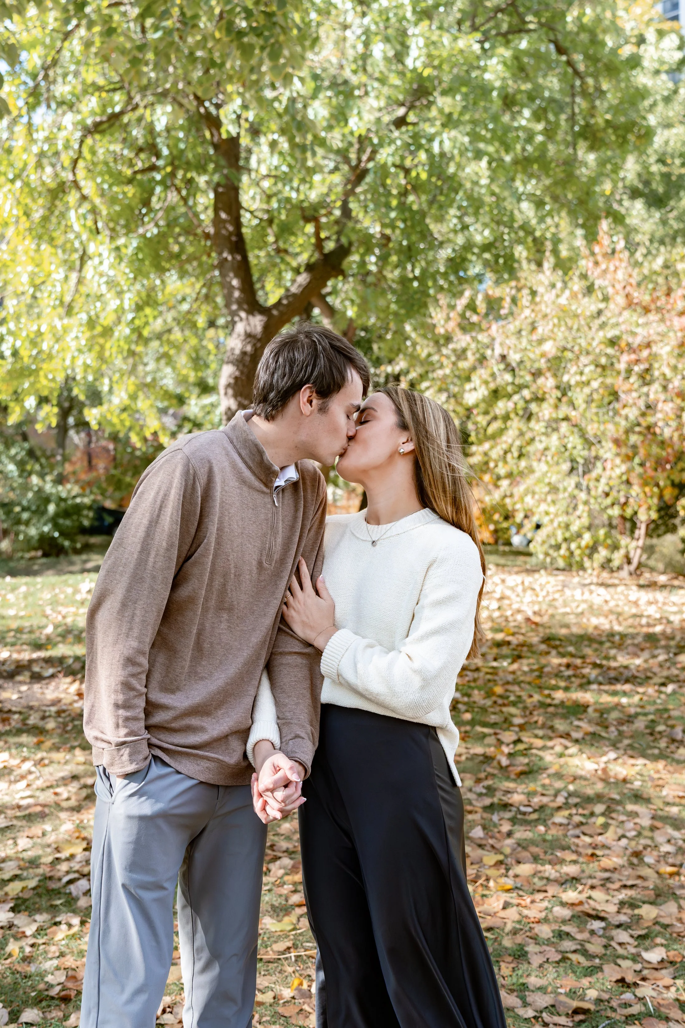 A young couple is kissing outdoors in a park with fall foliage.