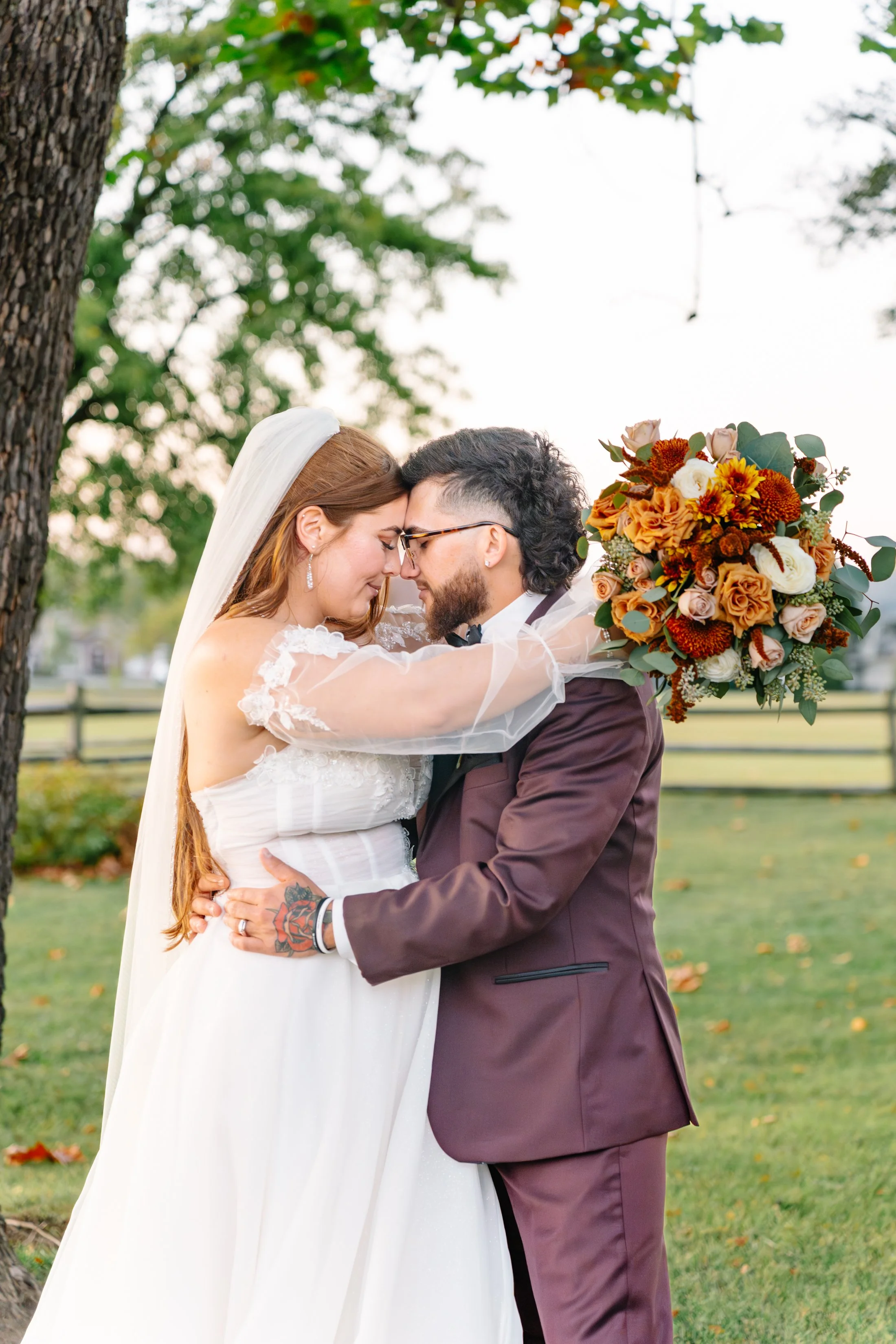 Bride and groom embrace outdoors, foreheads touching, holding each other. The bride wears a white wedding dress with lace details, and a veil, the groom wears a maroon suit with glasses and a beard. The bride holds a vibrant bouquet of orange, red, w