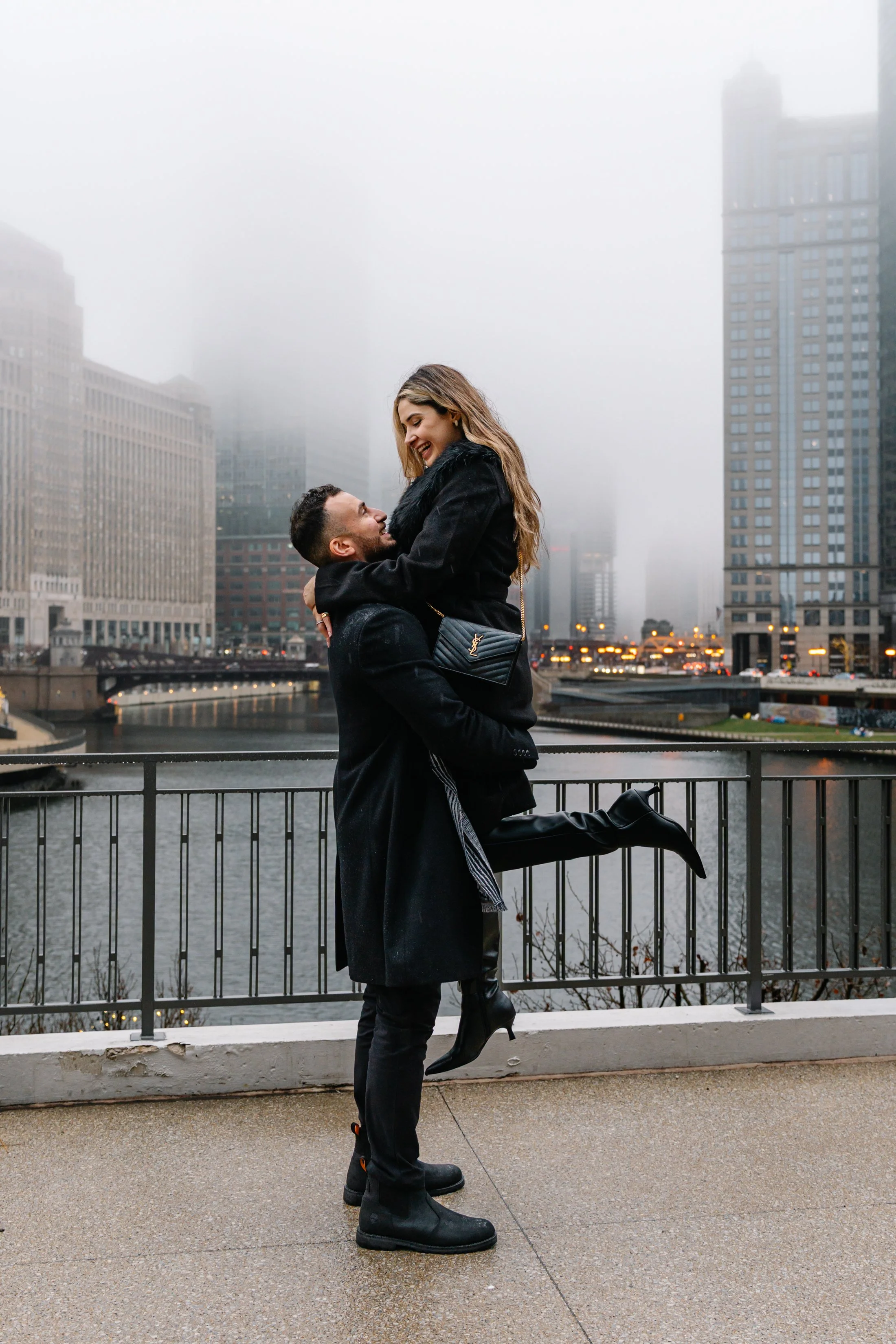 A couple embracing and smiling on a bridge in an urban setting with foggy city buildings in the background.