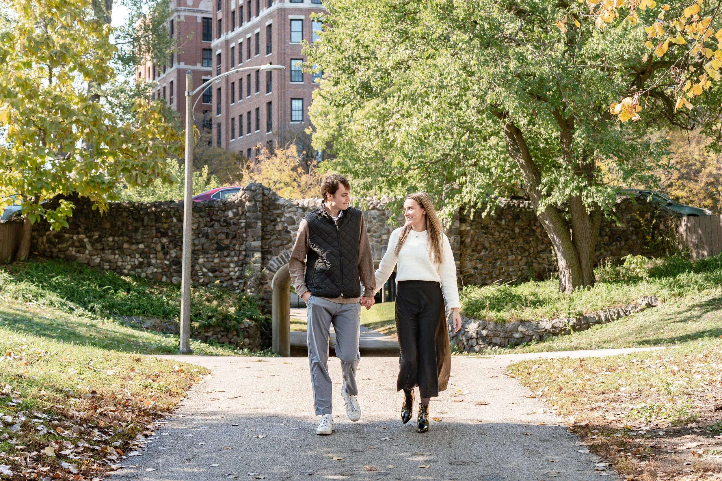 A young couple walking hand in hand on a sidewalk in a park, surrounded by autumn leaves and green trees, with a stone wall and residential buildings in the background.
