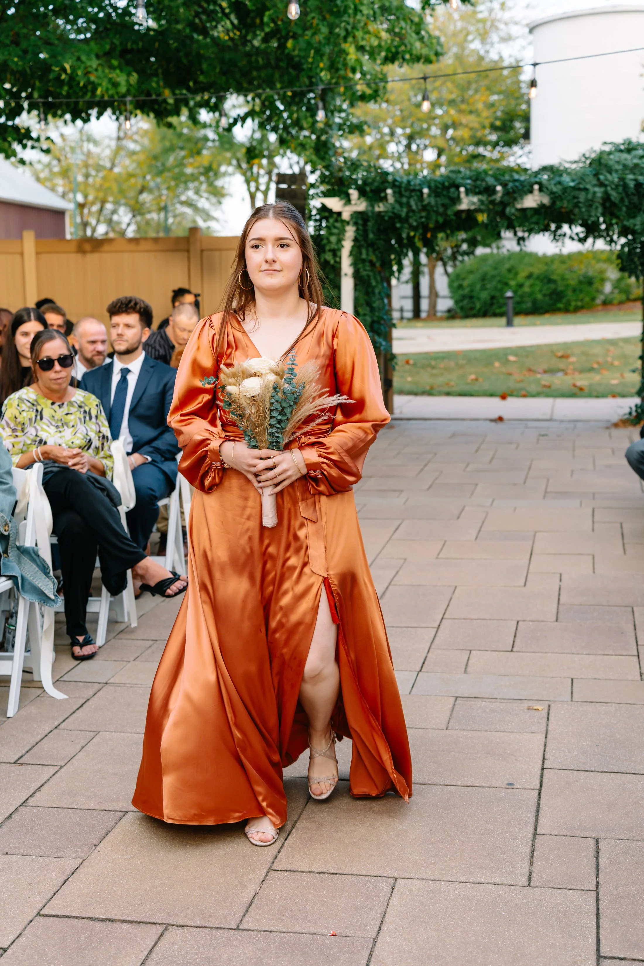 A young woman in a long satin burnt orange dress holds a bouquet of dried flowers as she walks down an outdoor aisle during a wedding ceremony. She is surrounded by seated guests, some dressed in formal attire, with a wooden fence, trees, and string 