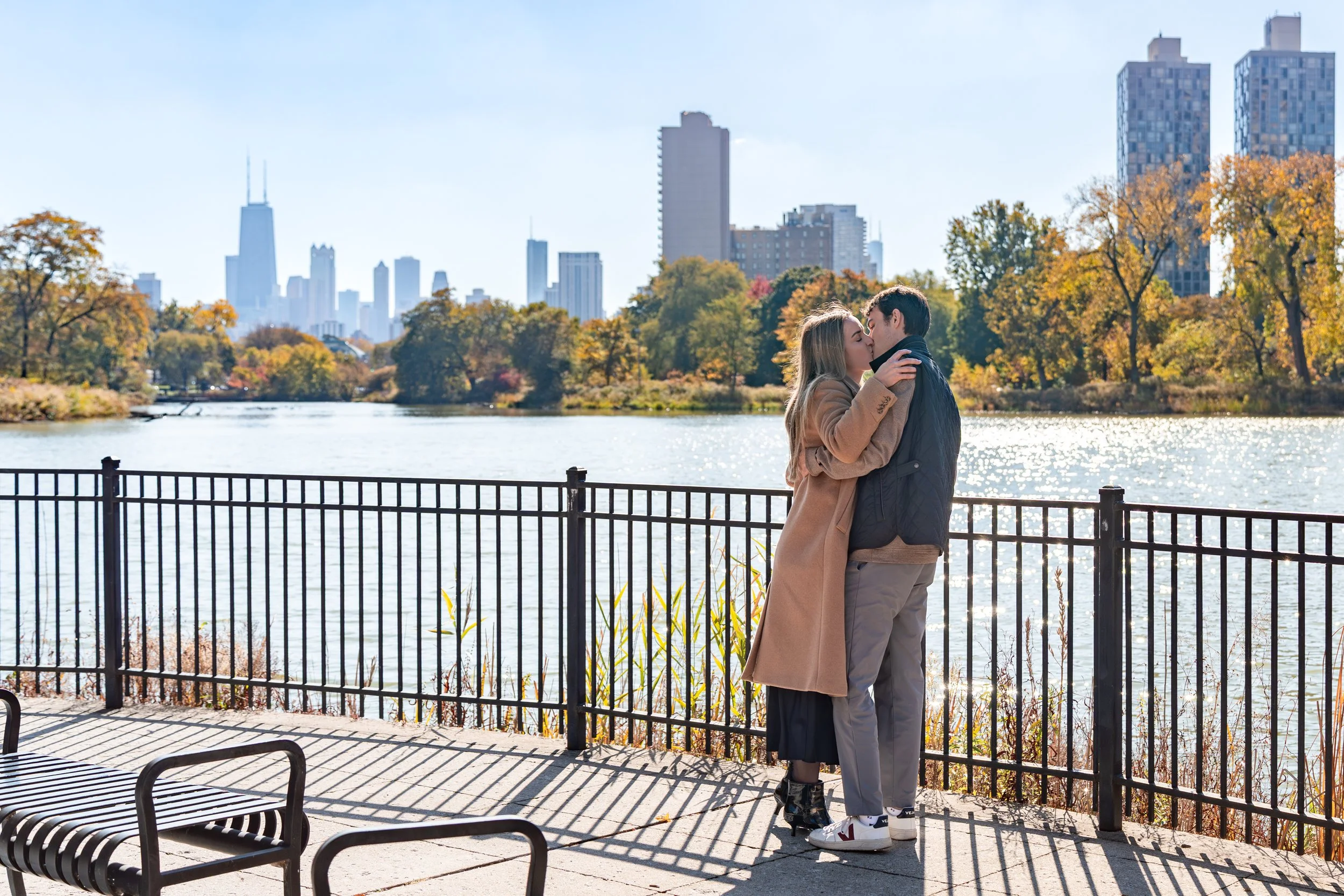 A couple kissing by a lakeside fence with city skyscrapers in the background on a sunny autumn day.