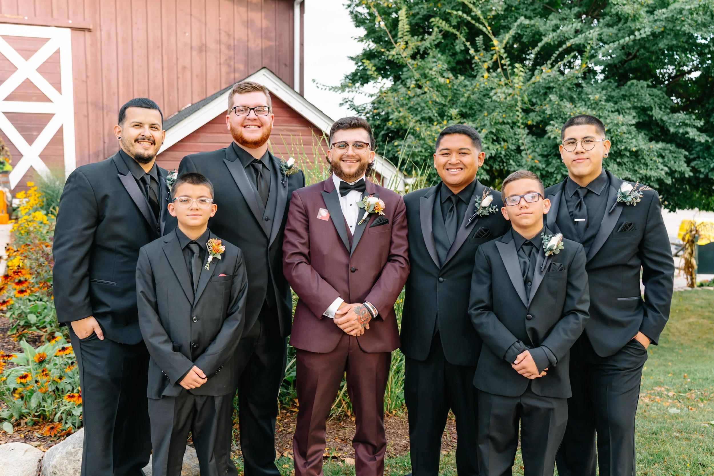 Group of seven men and boys dressed in formal suits, standing outdoors with a barn and trees in the background, smiling for a photo.