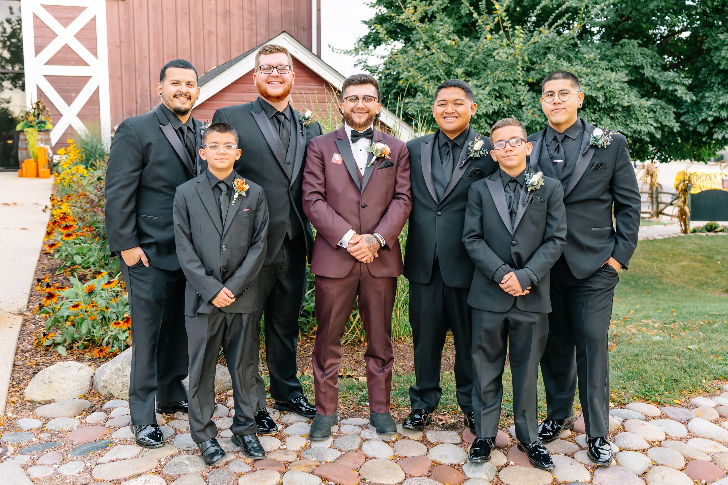 Group of seven men in tuxedos standing outdoors on a cobblestone path with fall flowers and greenery in the background.