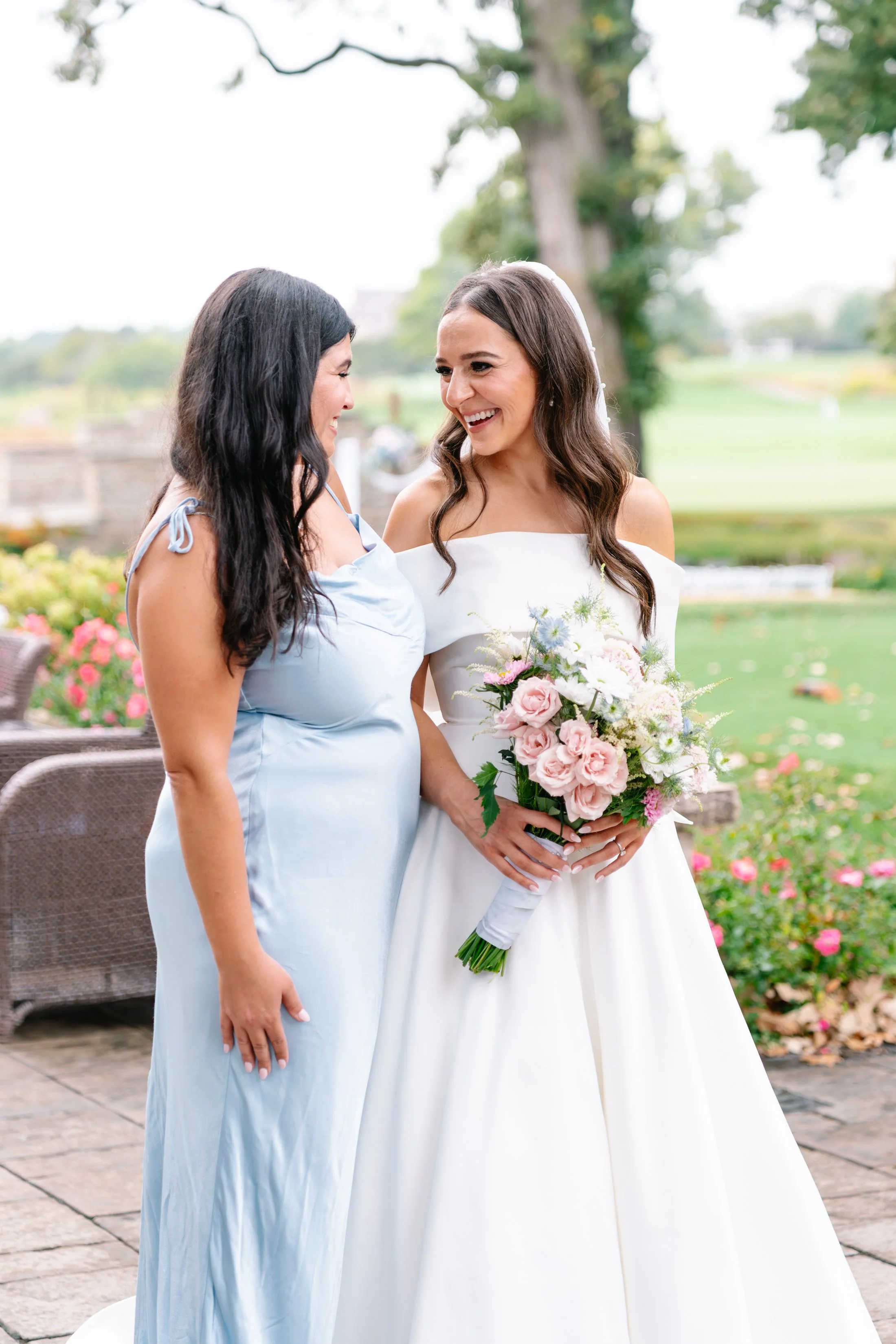 A bride in a white wedding dress holding a bouquet of pink and white flowers, smiling at a woman in a light blue dress, outside in a garden setting with greenery and pink roses.