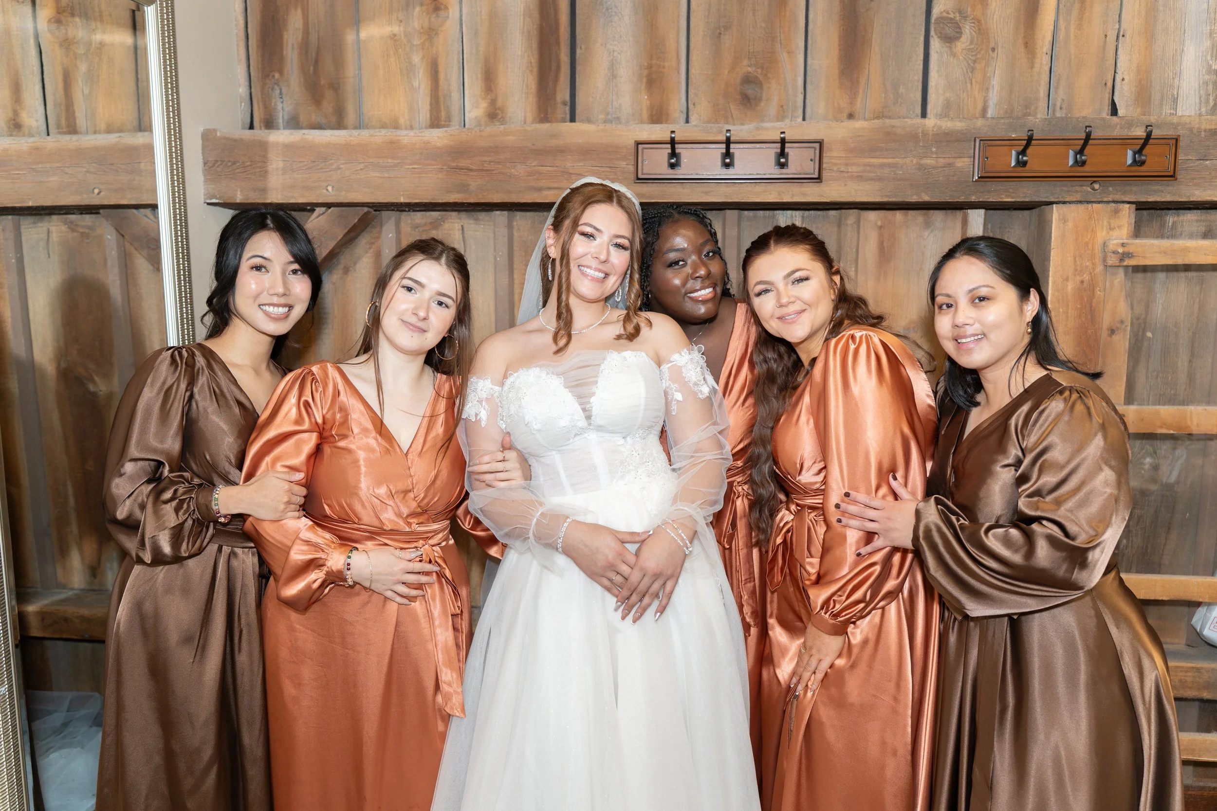 A bride in a white wedding dress surrounded by five bridesmaids in satin dresses in shades of brown and copper, standing in front of a wooden wall.