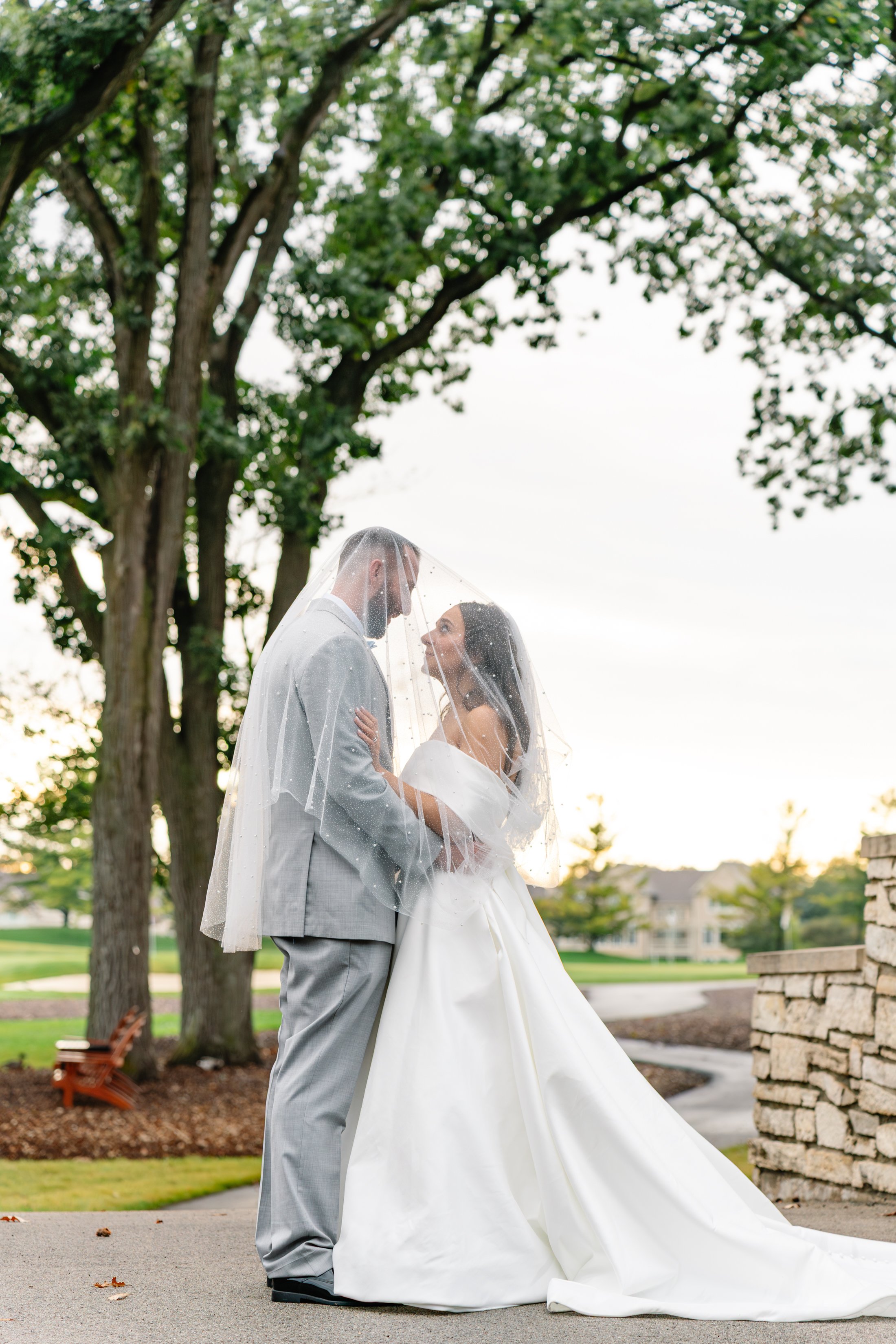 A bride and groom standing under a veil with a tree and a park background at sunset.