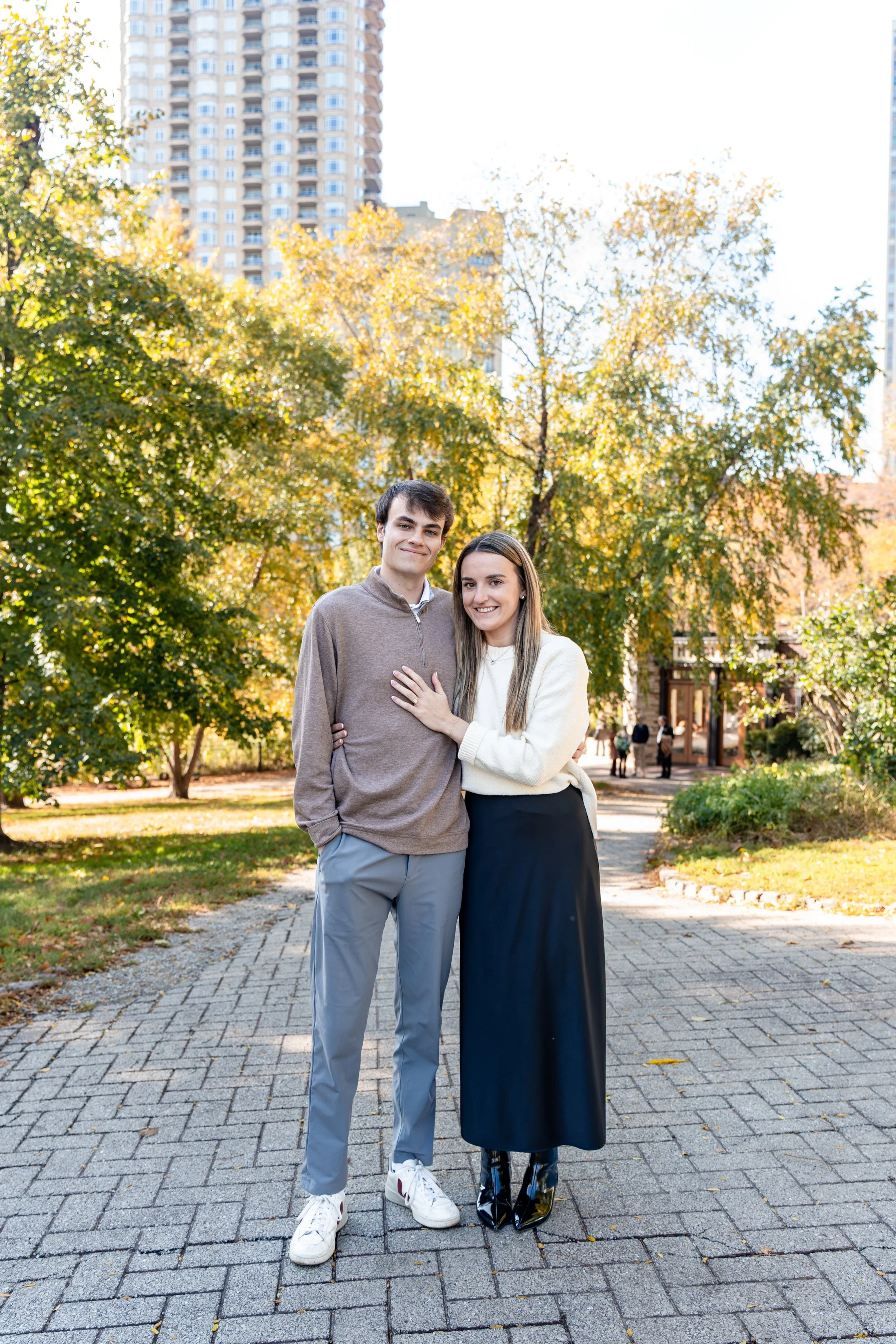 A young couple posing outdoors on a city park pathway during autumn, with tall buildings in the background.
