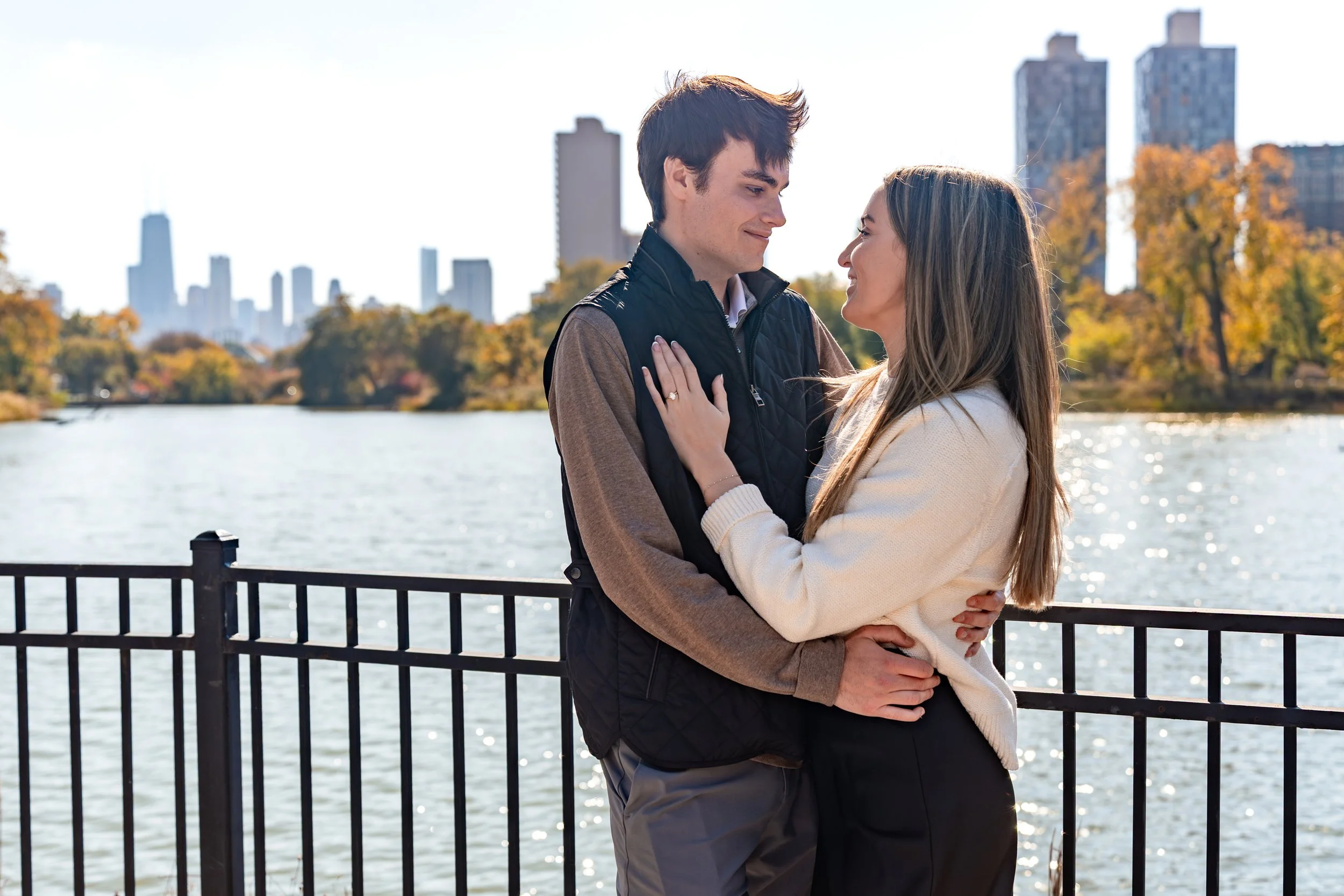 A young couple standing close together by a railing near a body of water, with a city skyline and trees in autumn colors in the background.