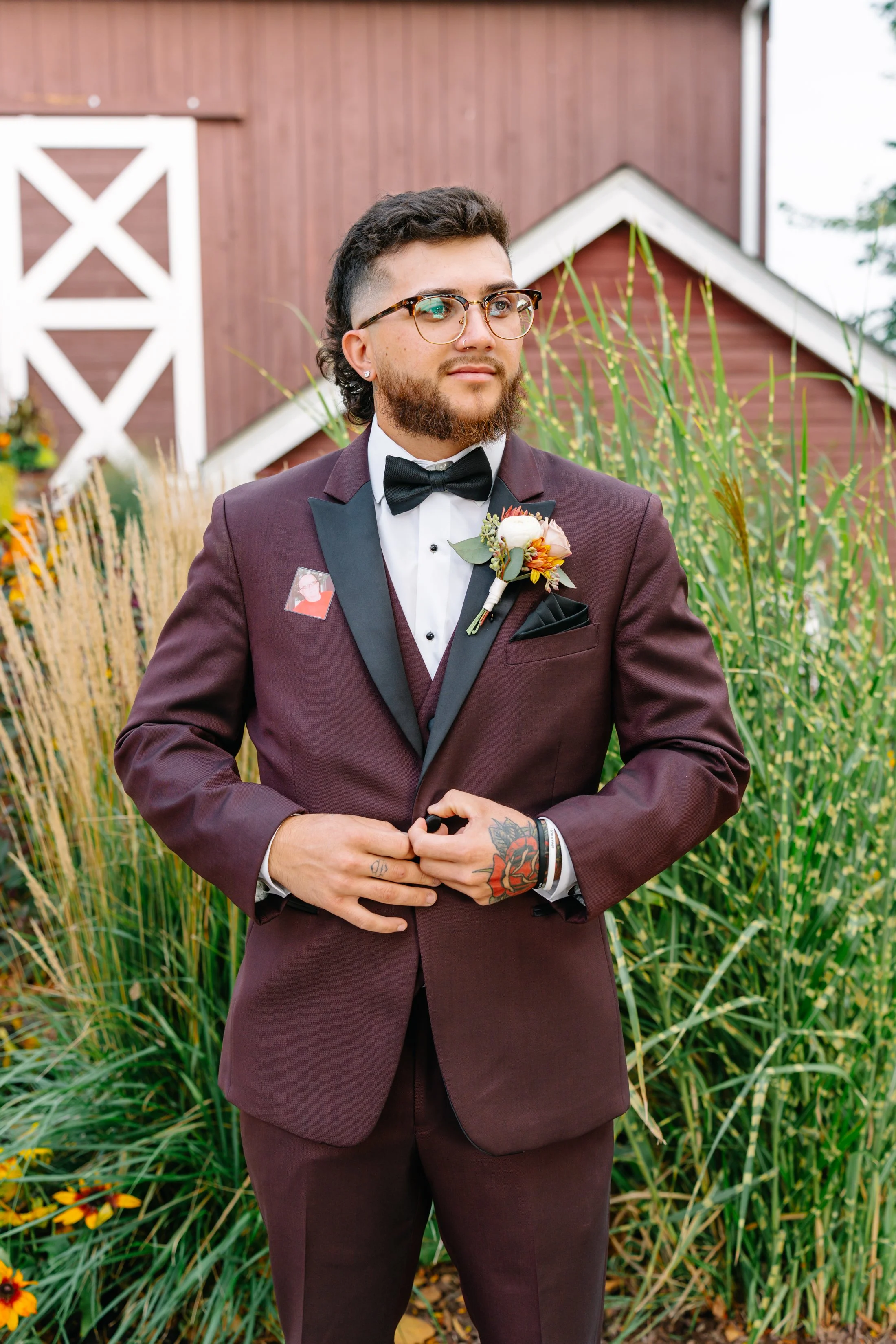 A man dressed in a burgundy tuxedo with black lapels, a white shirt, and a black bowtie, standing outdoors in front of green plants and a red wooden building, holding his hands near his waist.
