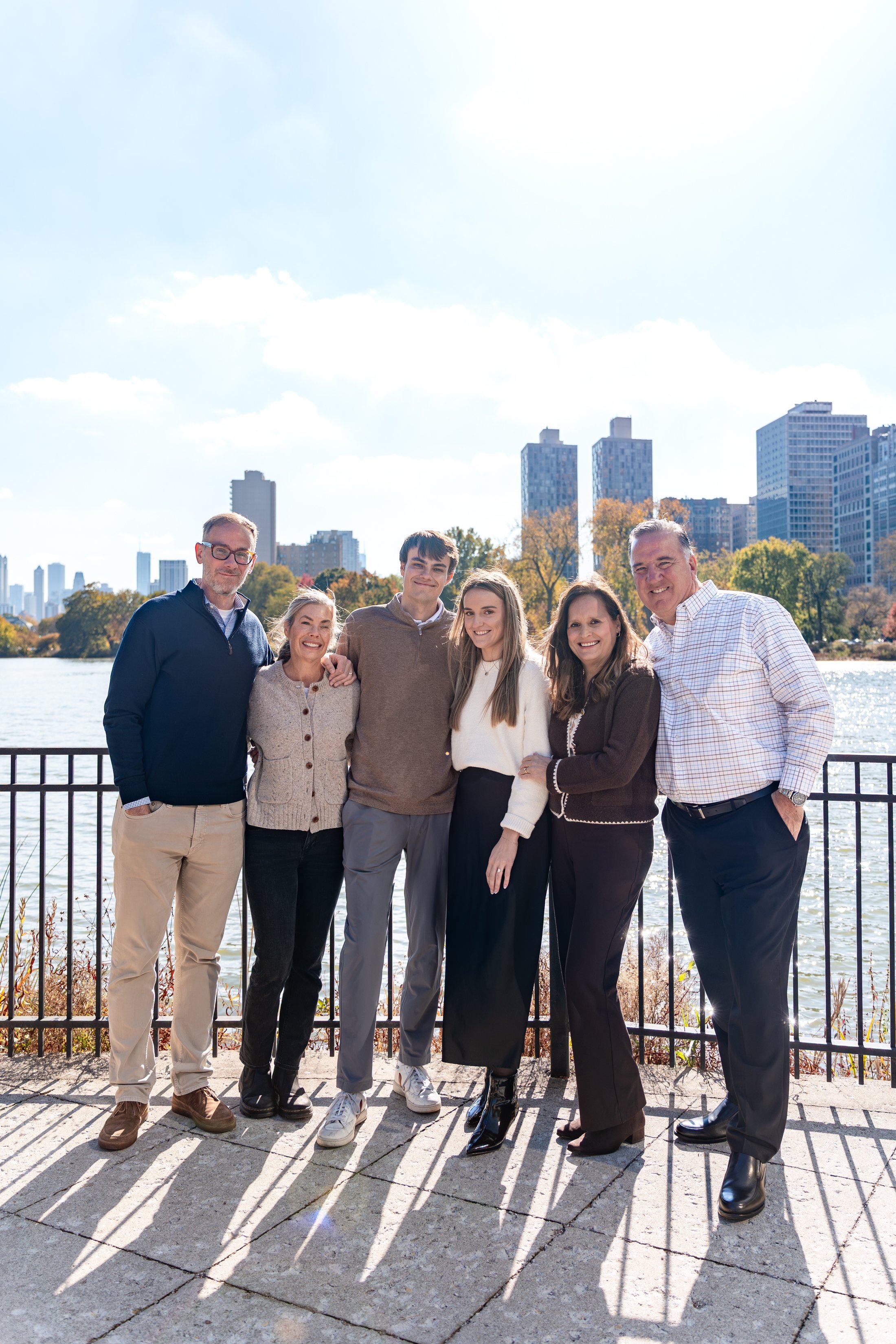 Group of six people standing outdoors near a fence with a city skyline and water in the background, smiling and posing for a photo.