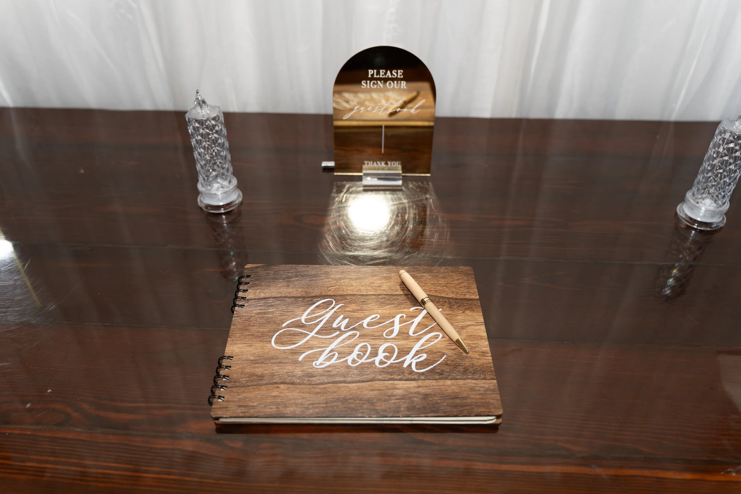 Guest book with a pen on a dark wooden table, flanked by two crystal glasses, and a small sign that reads, 'Please sign our guestbook, thank you.'