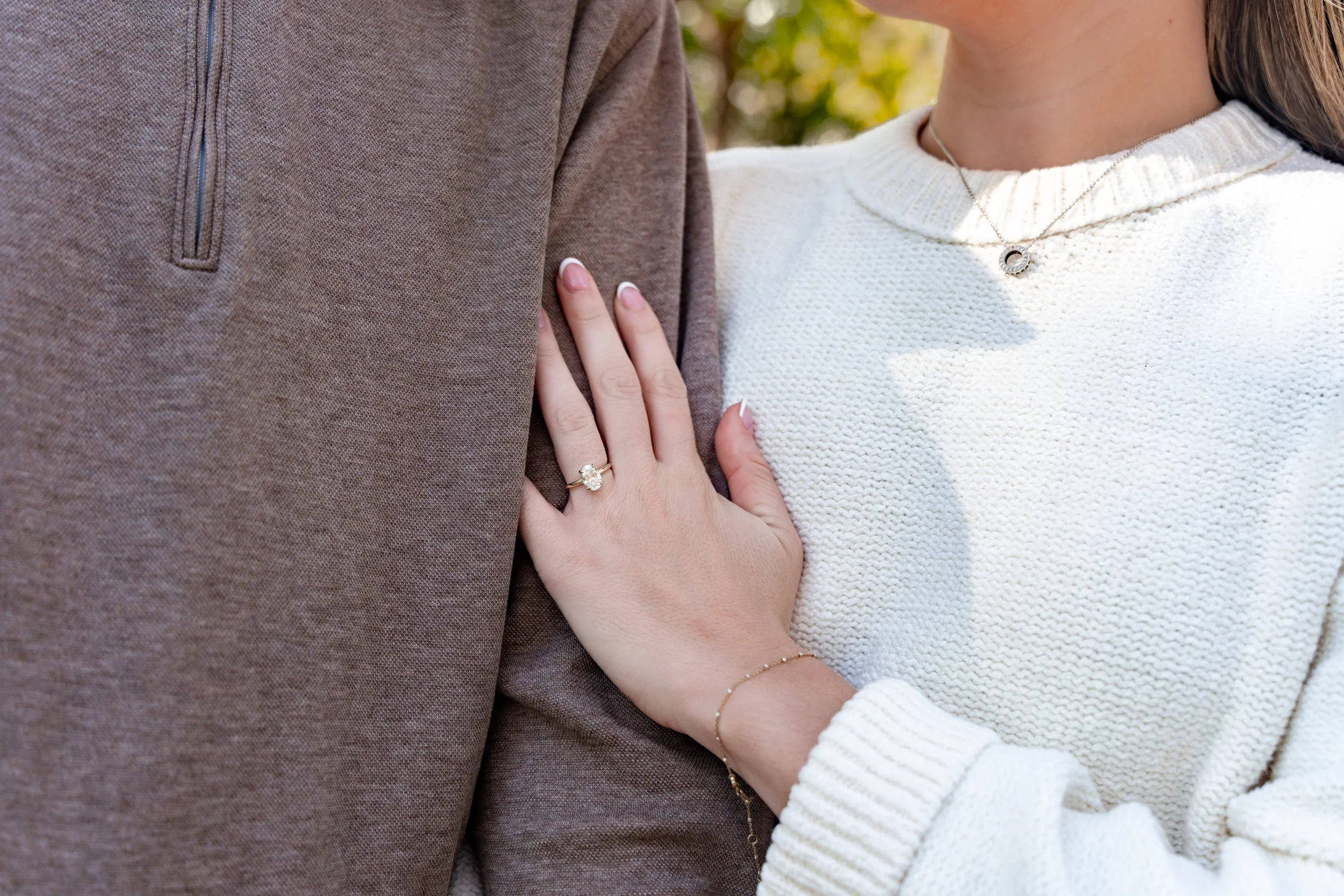 A woman gently holding a man's arm, showing her wedding ring, wearing a white sweater, and a delicate bracelet, with another necklace visible around her neck.