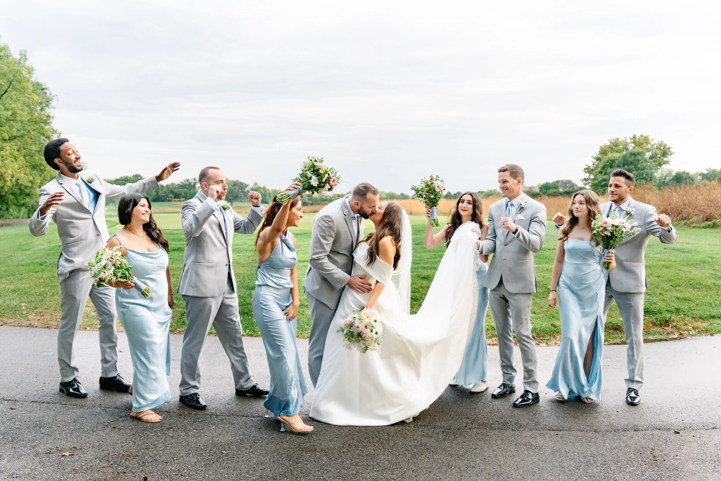 A wedding party outdoors, with the bride and groom kissing, surrounded by bridesmaids and groomsmen celebrating in suits and dresses.