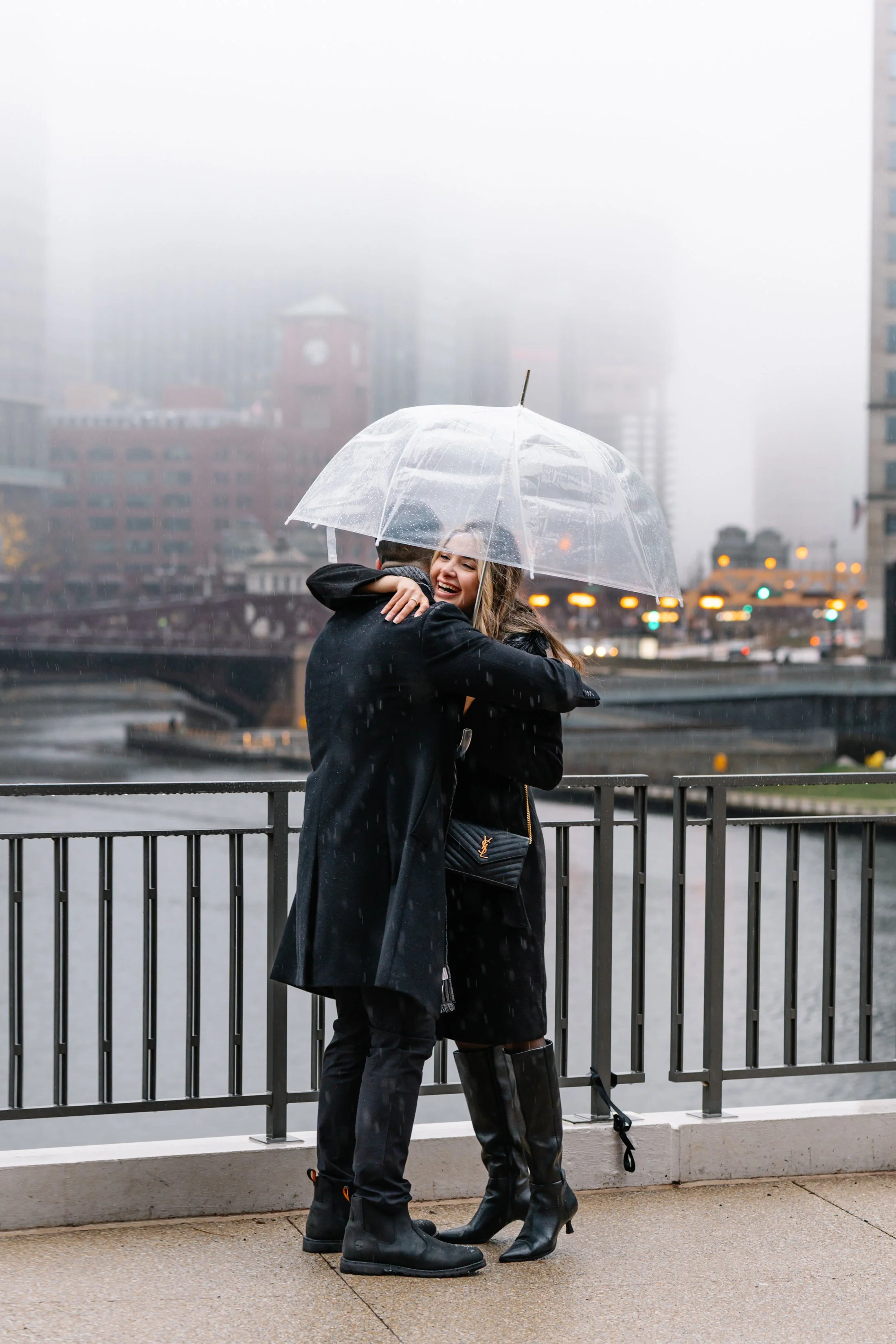 A man and woman embrace under a clear umbrella during rainy weather in an urban area, with buildings and a river in the background.