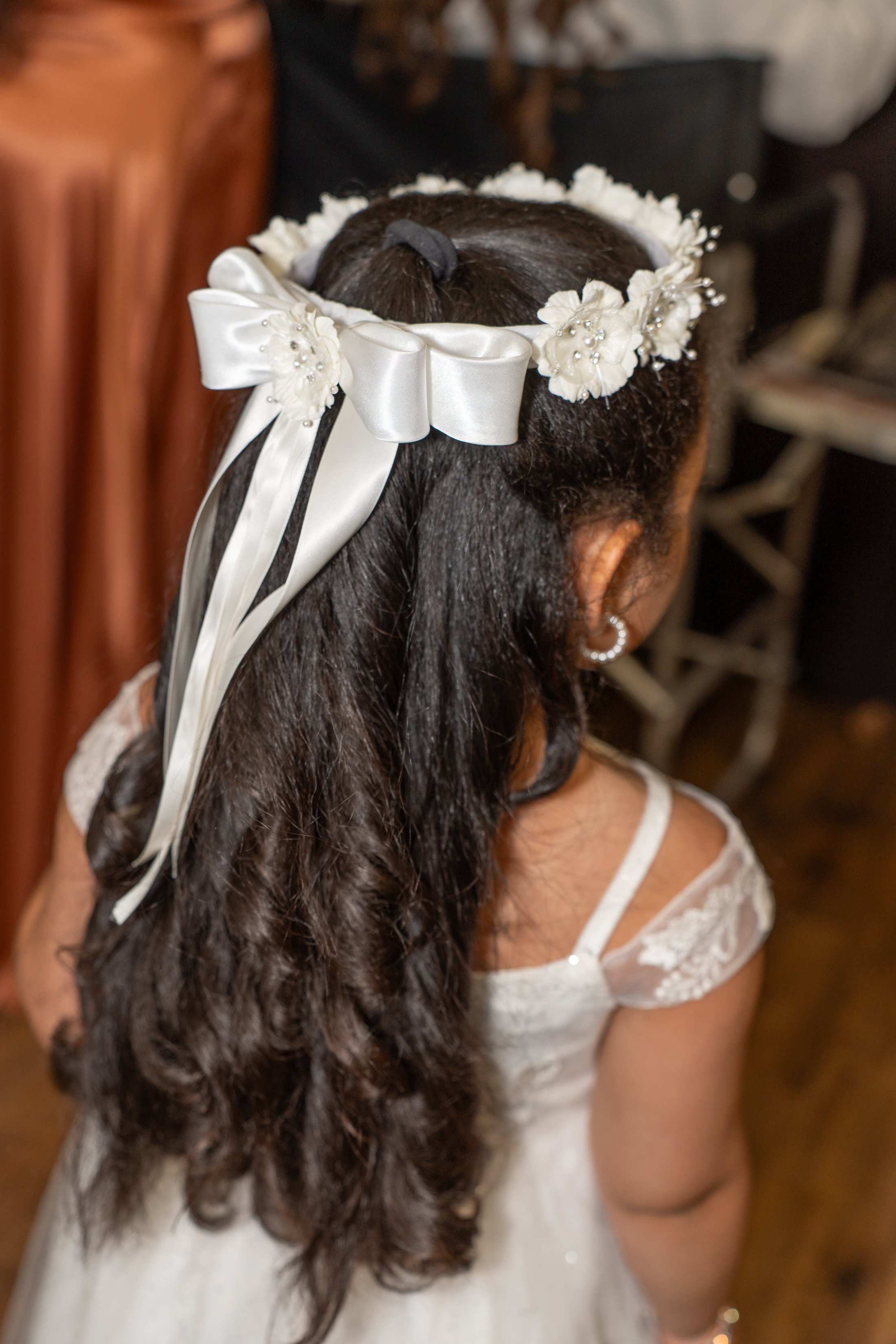 A young girl with long, dark curly hair wearing a floral headband with a large satin bow, pearls, and white fabric flowers. She has pearl earrings and a white lace dress with thin straps.