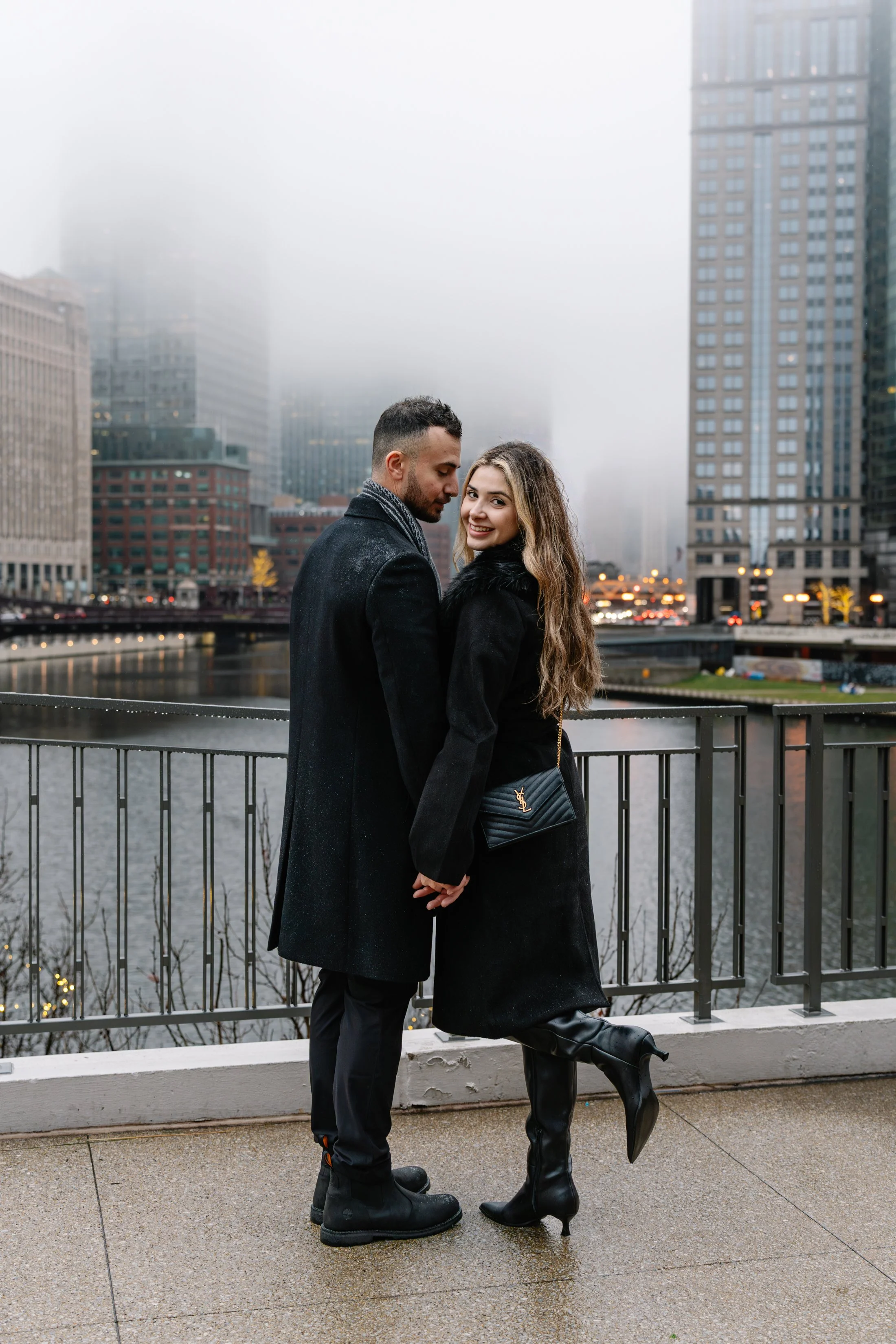 A couple standing close together by a metal railing overlooking a city river, with foggy skyscrapers in the background on a rainy day.