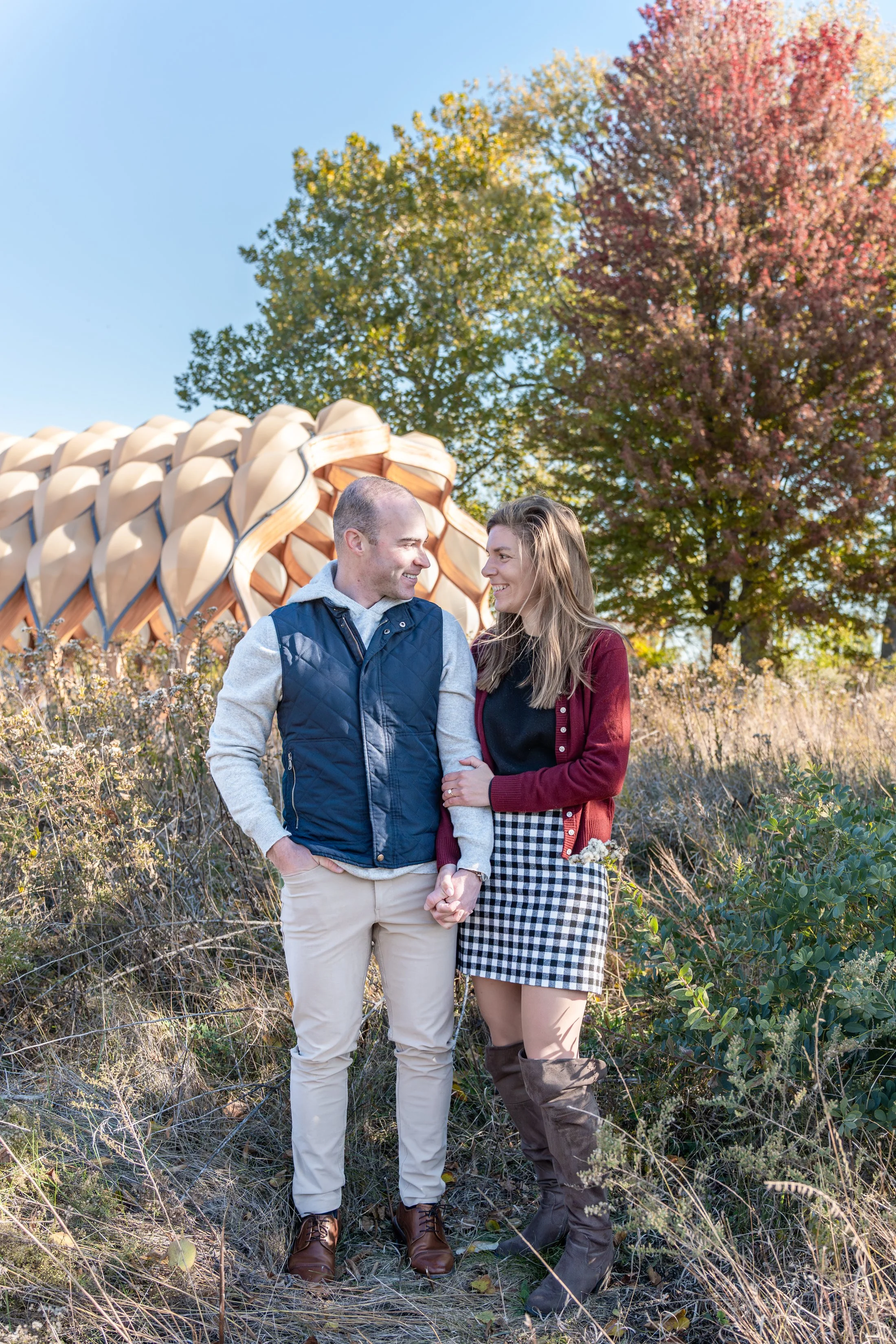A smiling couple holding hands outdoors on a sunny day, standing in front of a large sculpture resembling an acorn, with trees displaying fall foliage in the background.