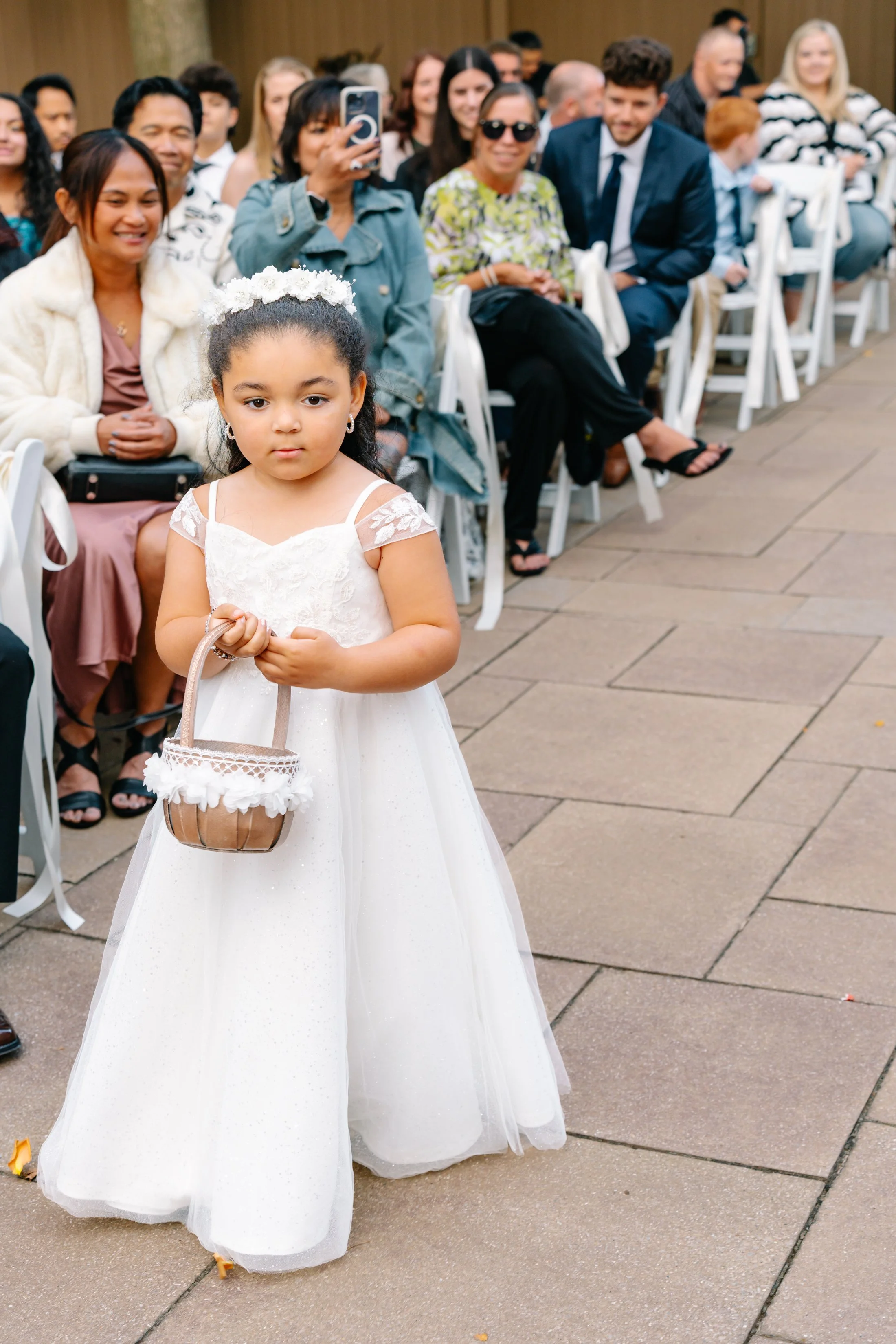 A young girl dressed in a white dress with lace details, holding a small basket, standing in front of seated guests at a wedding ceremony.
