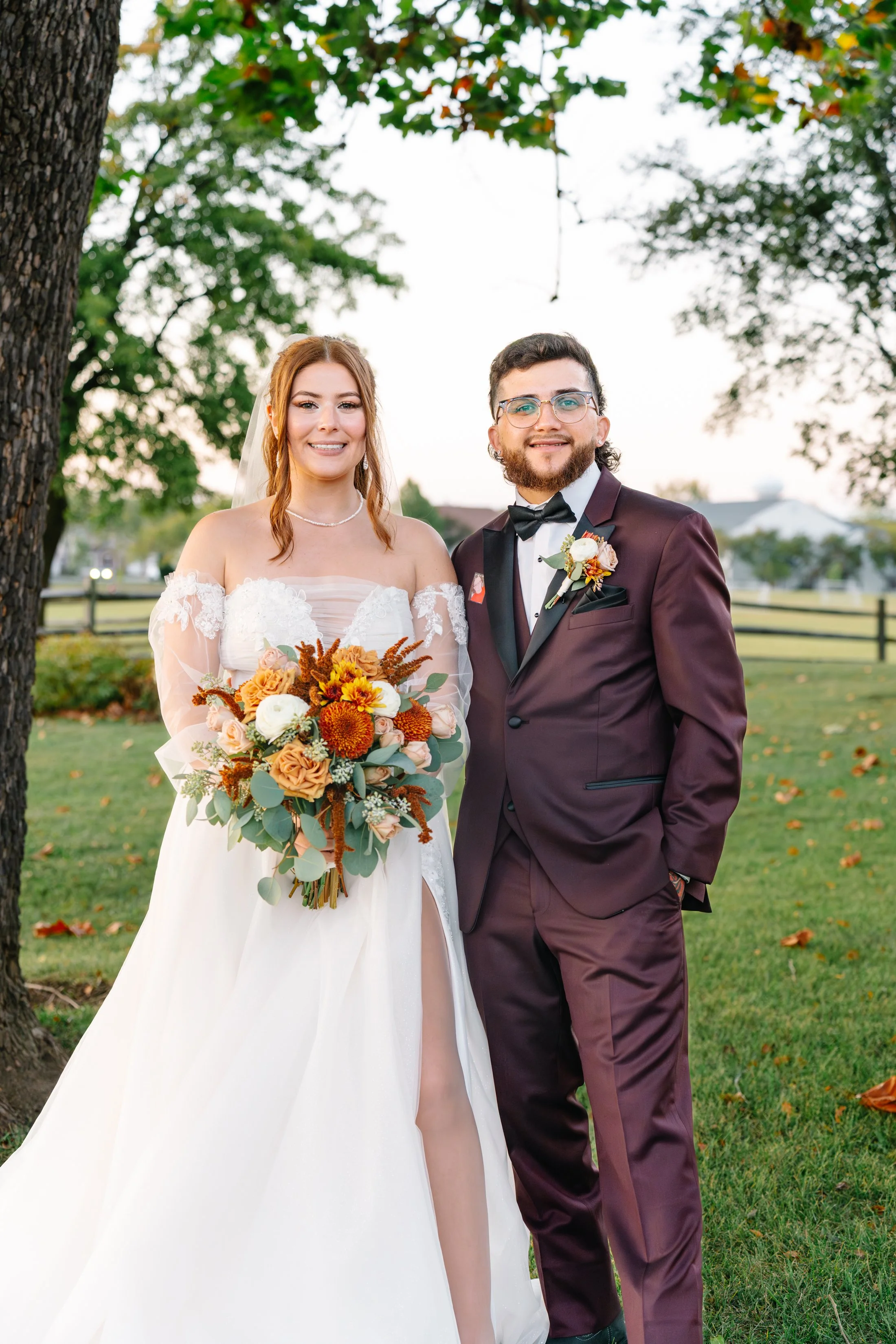 A bride and groom standing outdoors, smiling, with trees and a fence in the background. The bride is holding a bouquet of orange, white, and pink flowers, and is wearing a white wedding gown with lace details and a slit. The groom is dressed in a bur