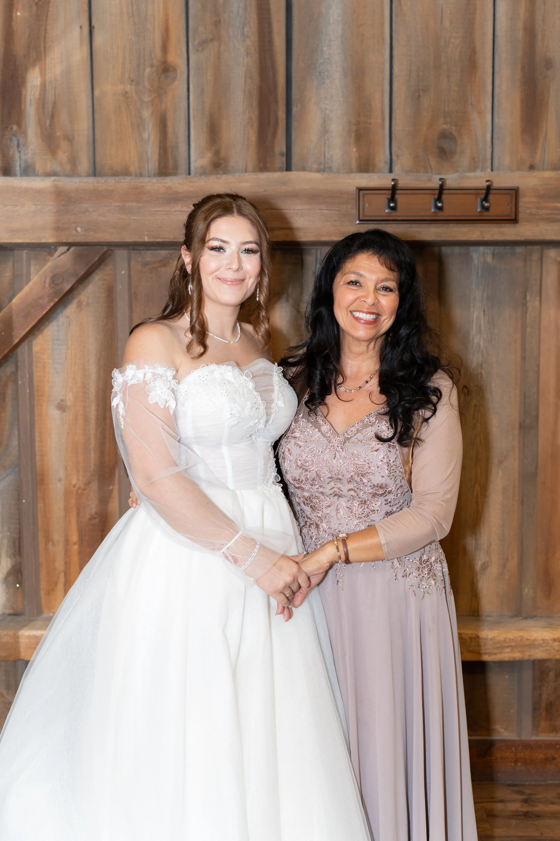A bride in a white wedding gown holding hands with a woman in a lavender dress, standing against a wooden wall.