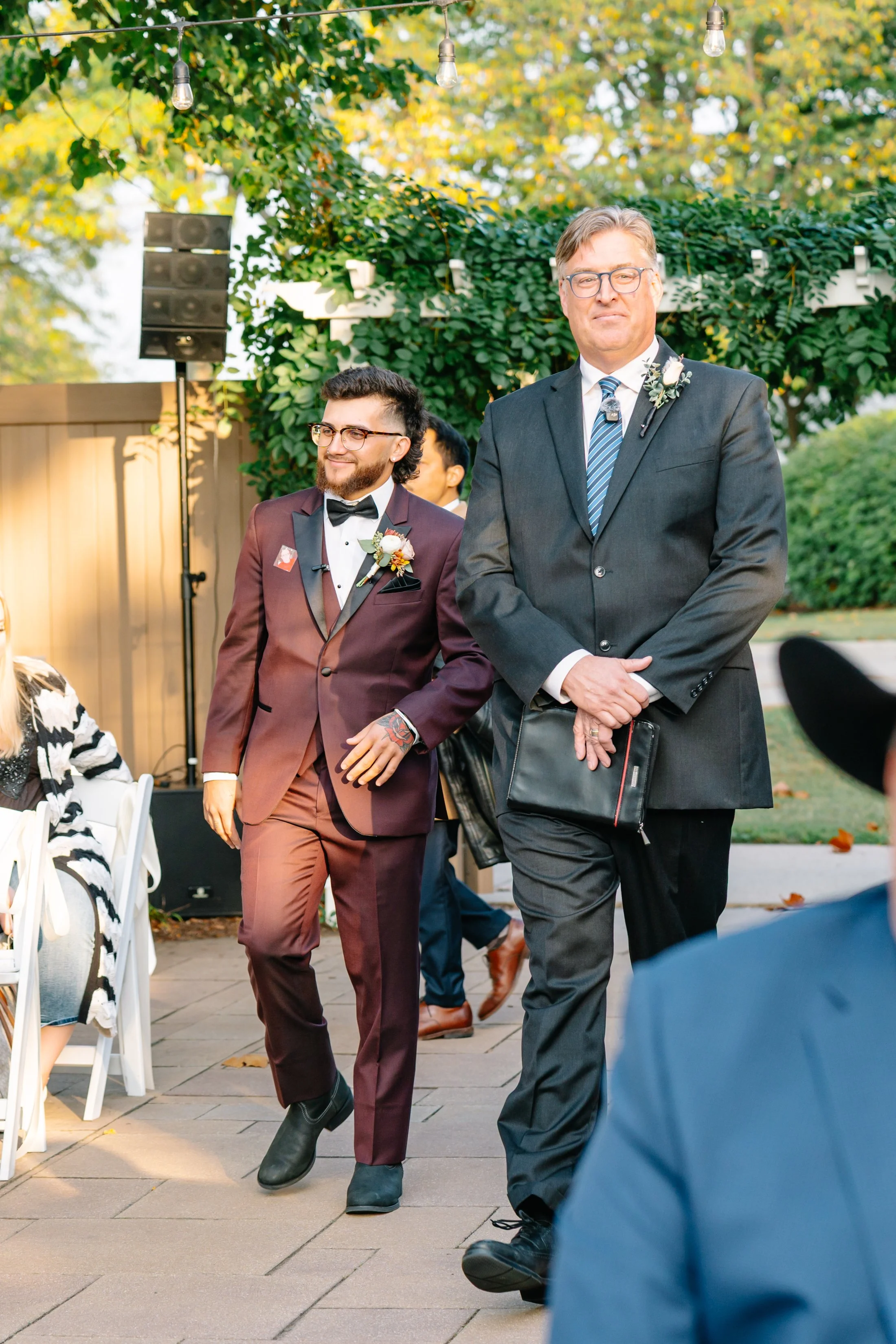 Two men, one in a dark gray suit and the other in a burgundy suit, walking at an outdoor event surrounded by greenery and string lights, possibly during a wedding or celebration.