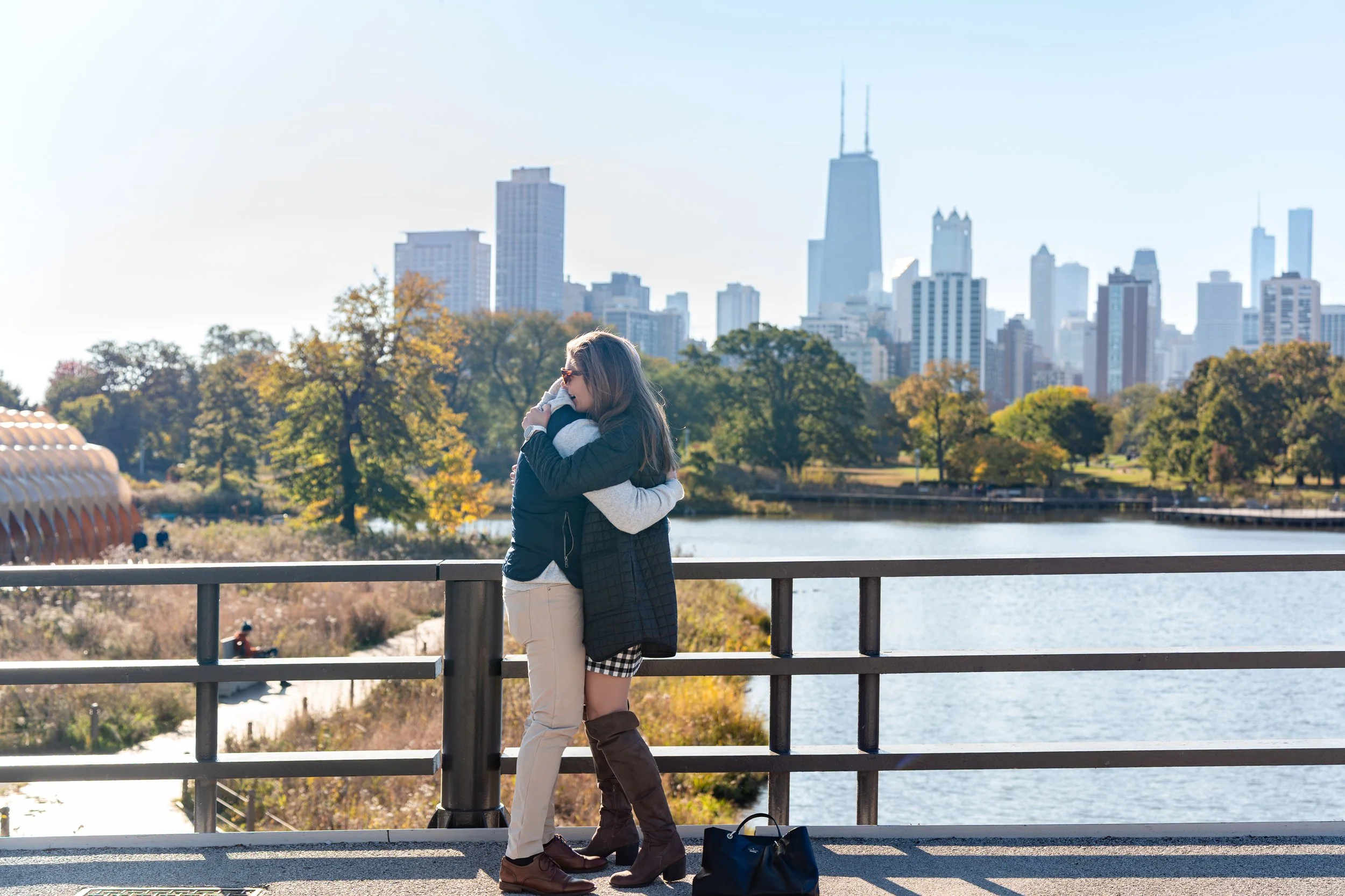 Two women hugging on a bridge overlooking a lake with the Chicago skyline in the background.