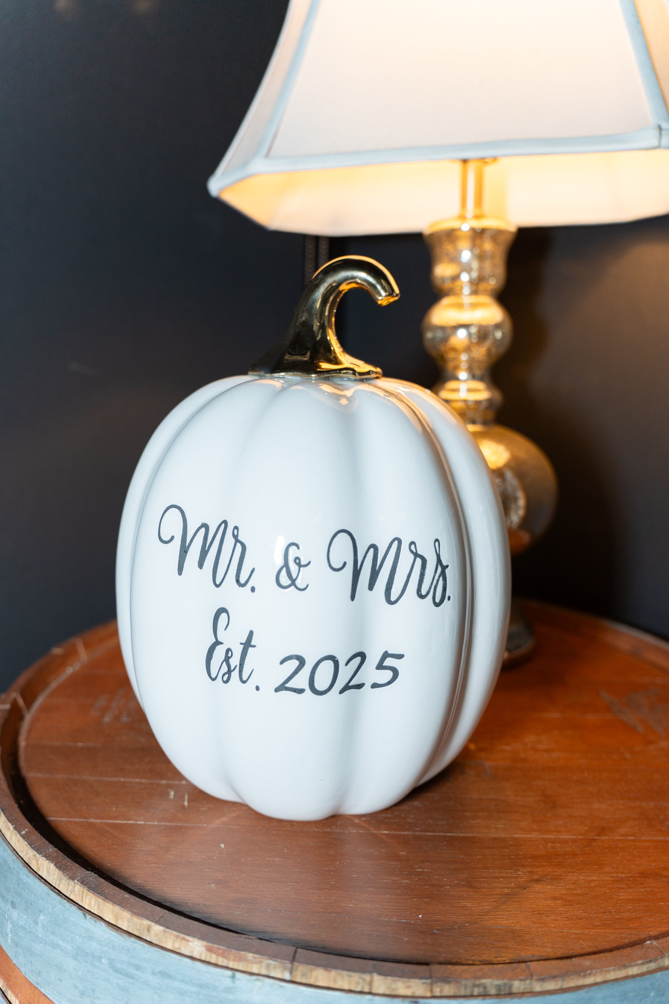 A white pumpkin-shaped decoration with gold stem, inscribed with 'Mr. & Mrs. Est. 2025,' placed on a wooden surface next to a table lamp with a white shade and gold base.
