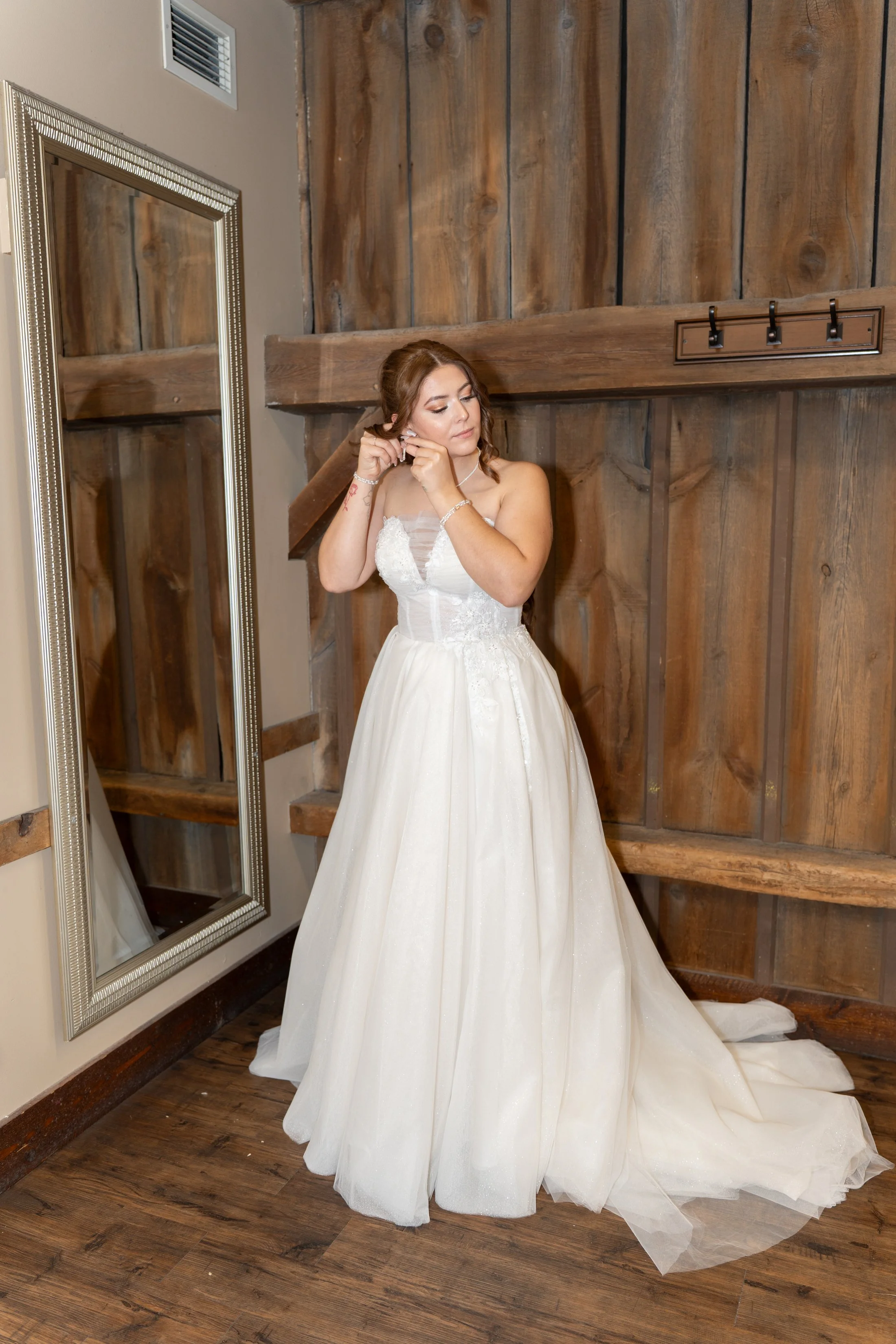 A woman in a white wedding dress standing and putting on earrings in front of a mirror with a wooden wall background.