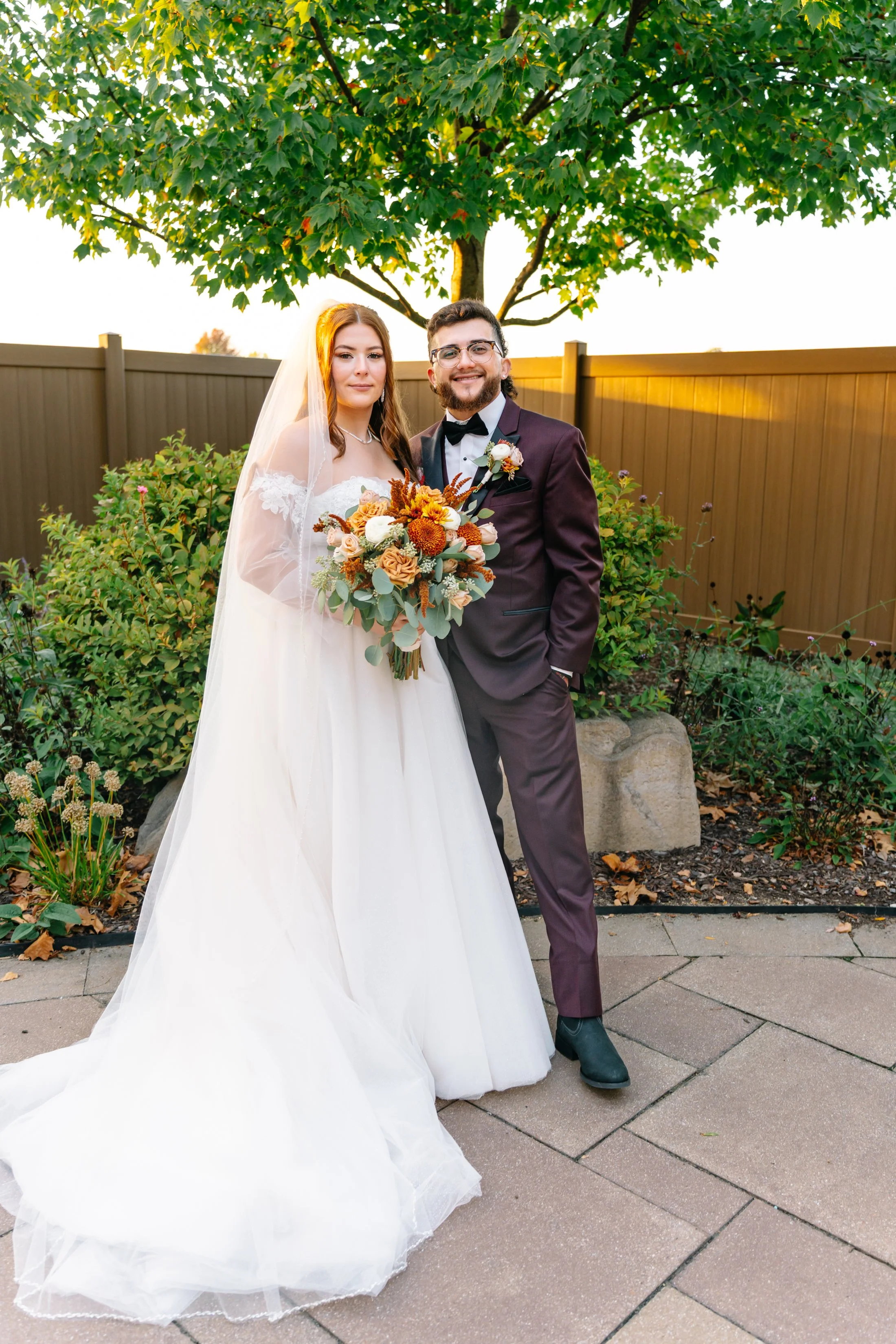 A bride and groom stand together outdoors, smiling, with the bride holding a bouquet of flowers. The bride is dressed in a white wedding gown with lace details, and the groom in a maroon tuxedo with a black bow tie. They are in a garden with green bu