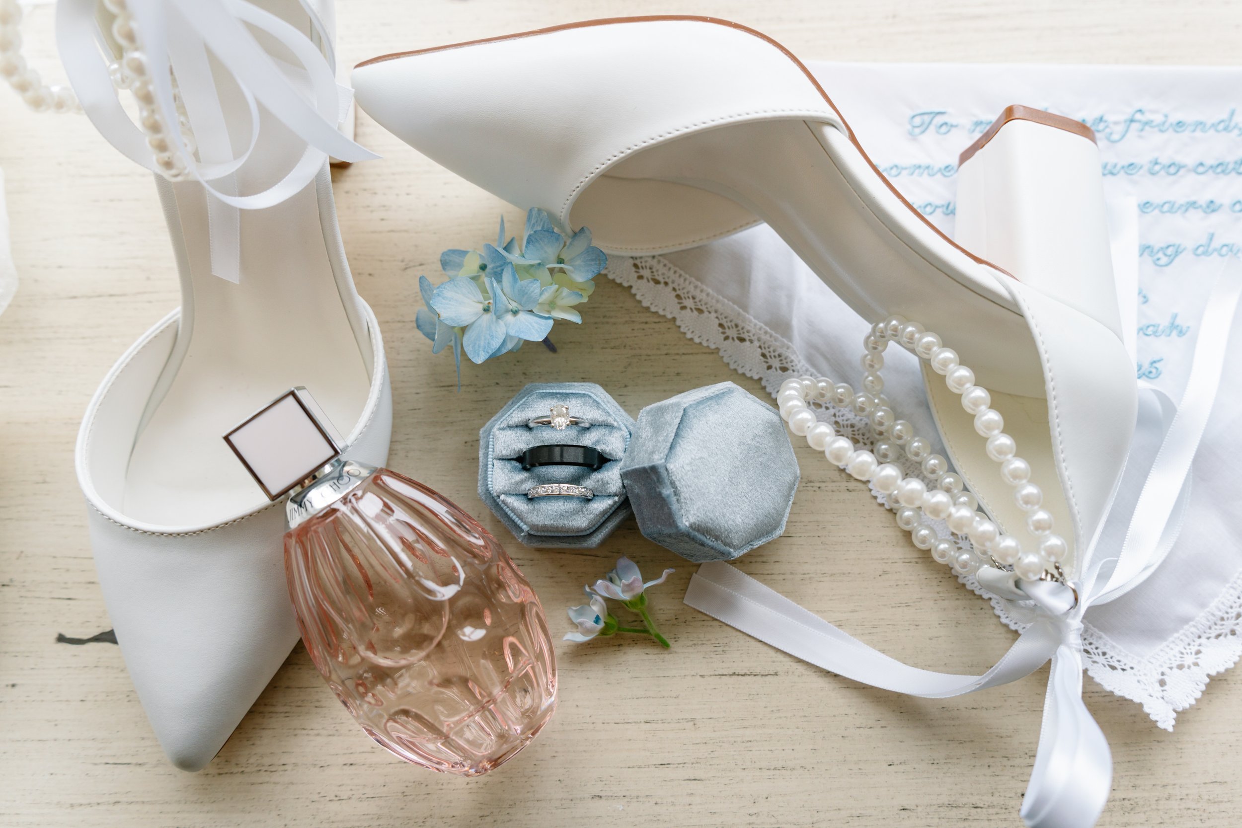 White high-heeled shoes, a pearl necklace, a perfume bottle, a blue floral hair accessory, and a ring in a ring box arranged on a light-colored wooden surface, with a white lace-trimmed cloth and a handwritten note in the background.
