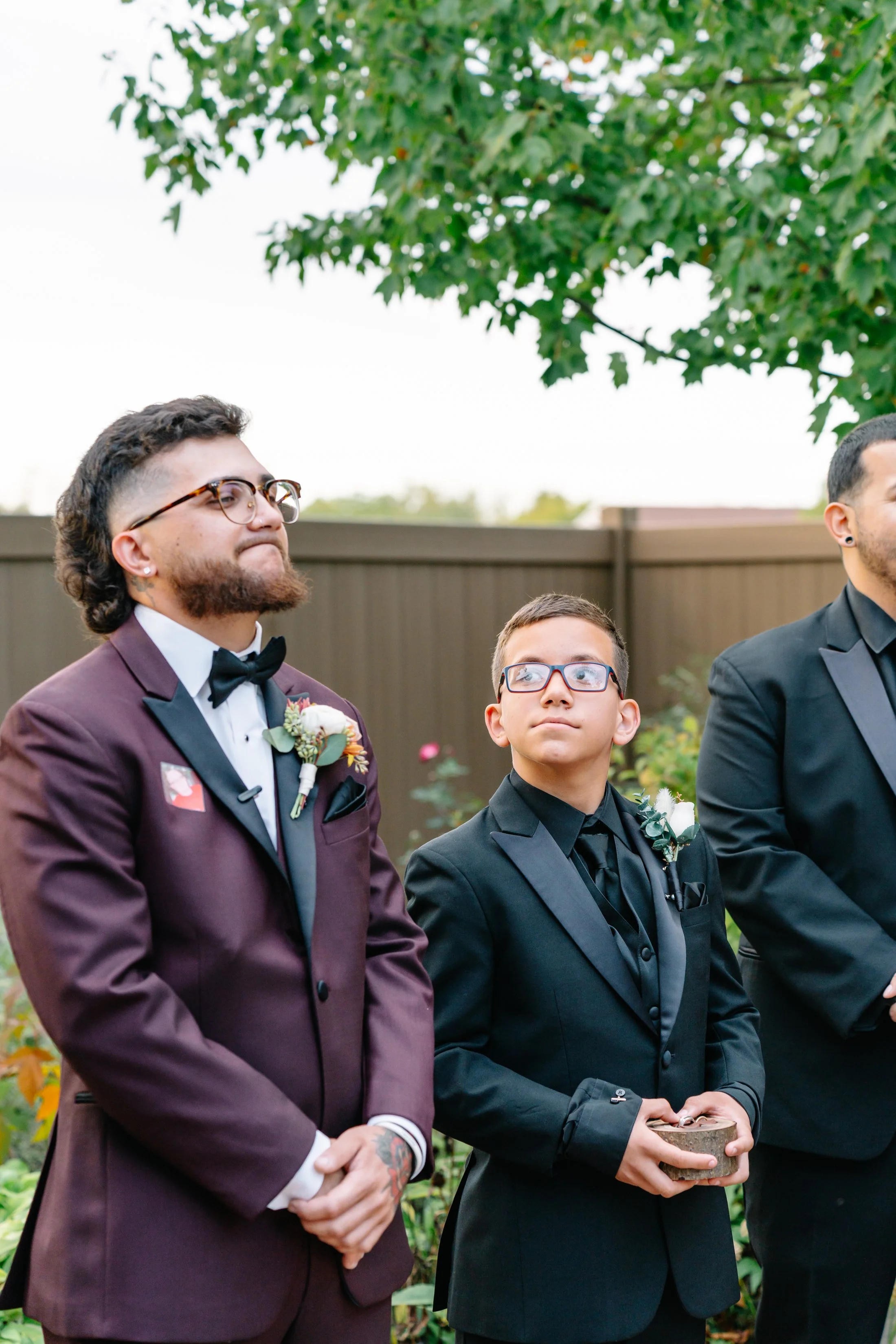 Three men in formal attire standing outdoors during daytime, with the two on the left wearing tuxedos and boutonnieres, at a garden or backyard event, possibly a wedding.