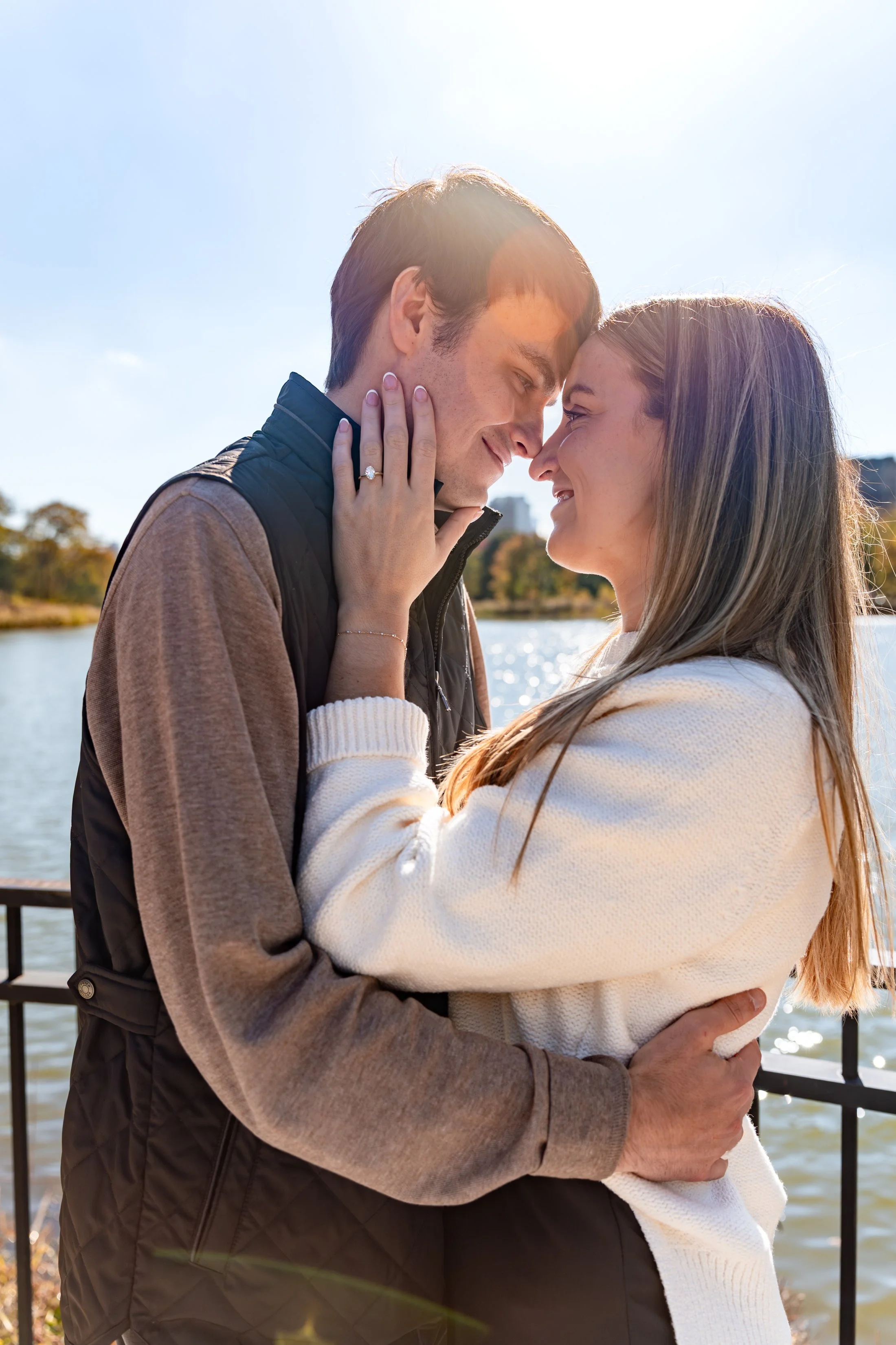 A smiling couple standing close together outdoors near a body of water with trees in the background, enjoying a sunny day.