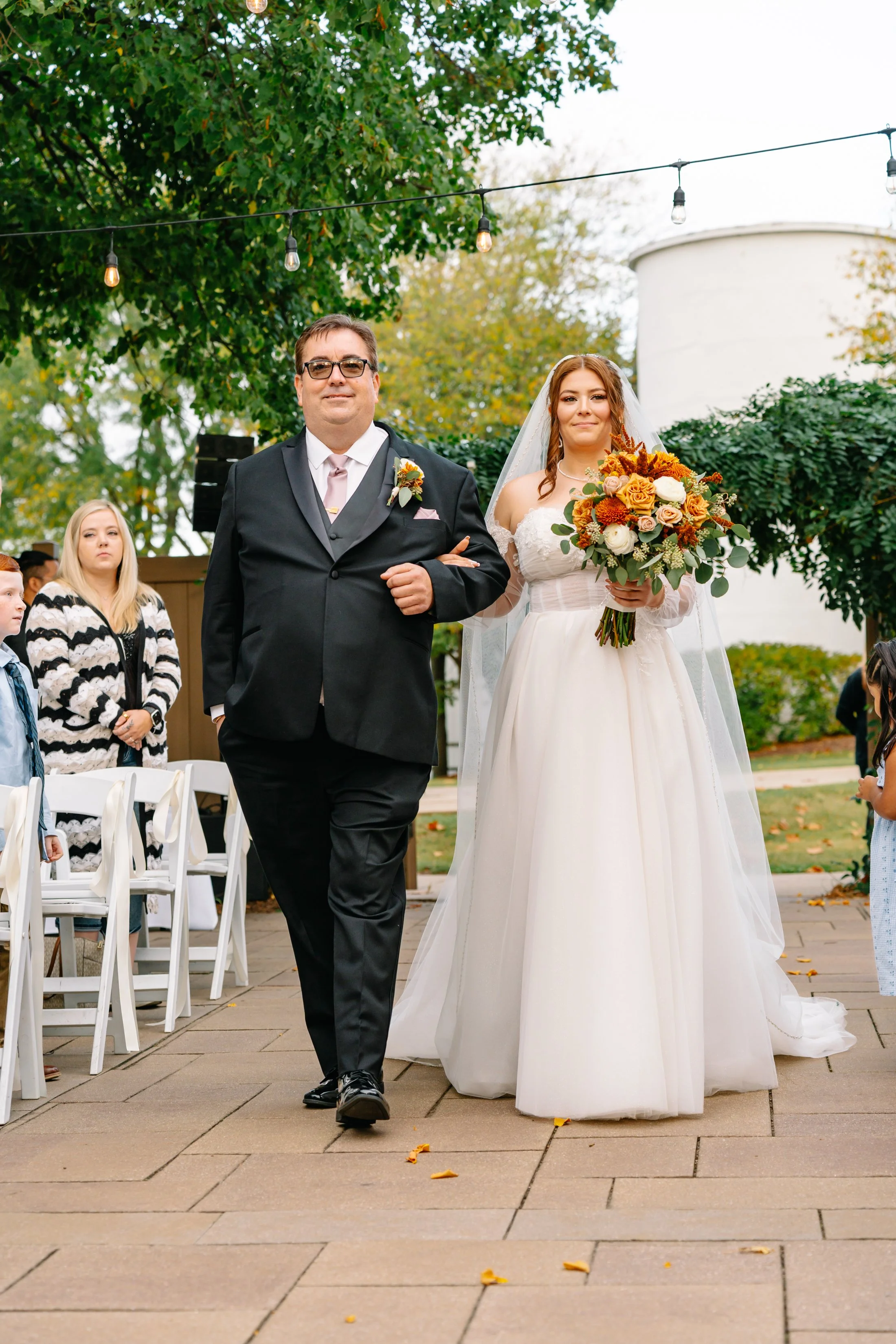 A bride walking down the aisle with her father at an outdoor wedding ceremony during daytime. The bride is wearing a white wedding gown and veil, holding a bouquet of orange, white, and yellow flowers, with trees and string lights in the background.