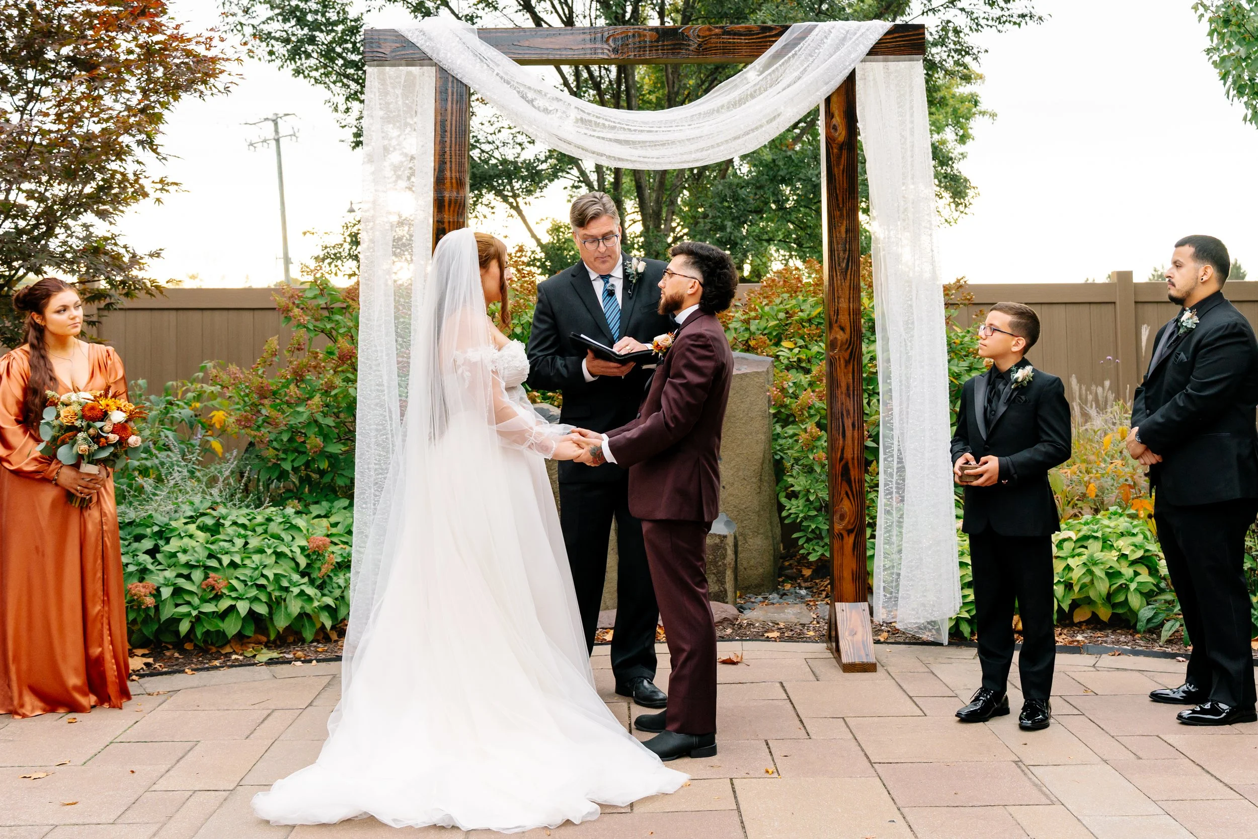 A wedding ceremony outdoors with a bride and groom holding hands under a wooden archway with white fabric, officiated by a man holding a book. Bridesmaid and groomsmen stand nearby, the bridesmaid holding a bouquet, all dressed in formal attire. Gree