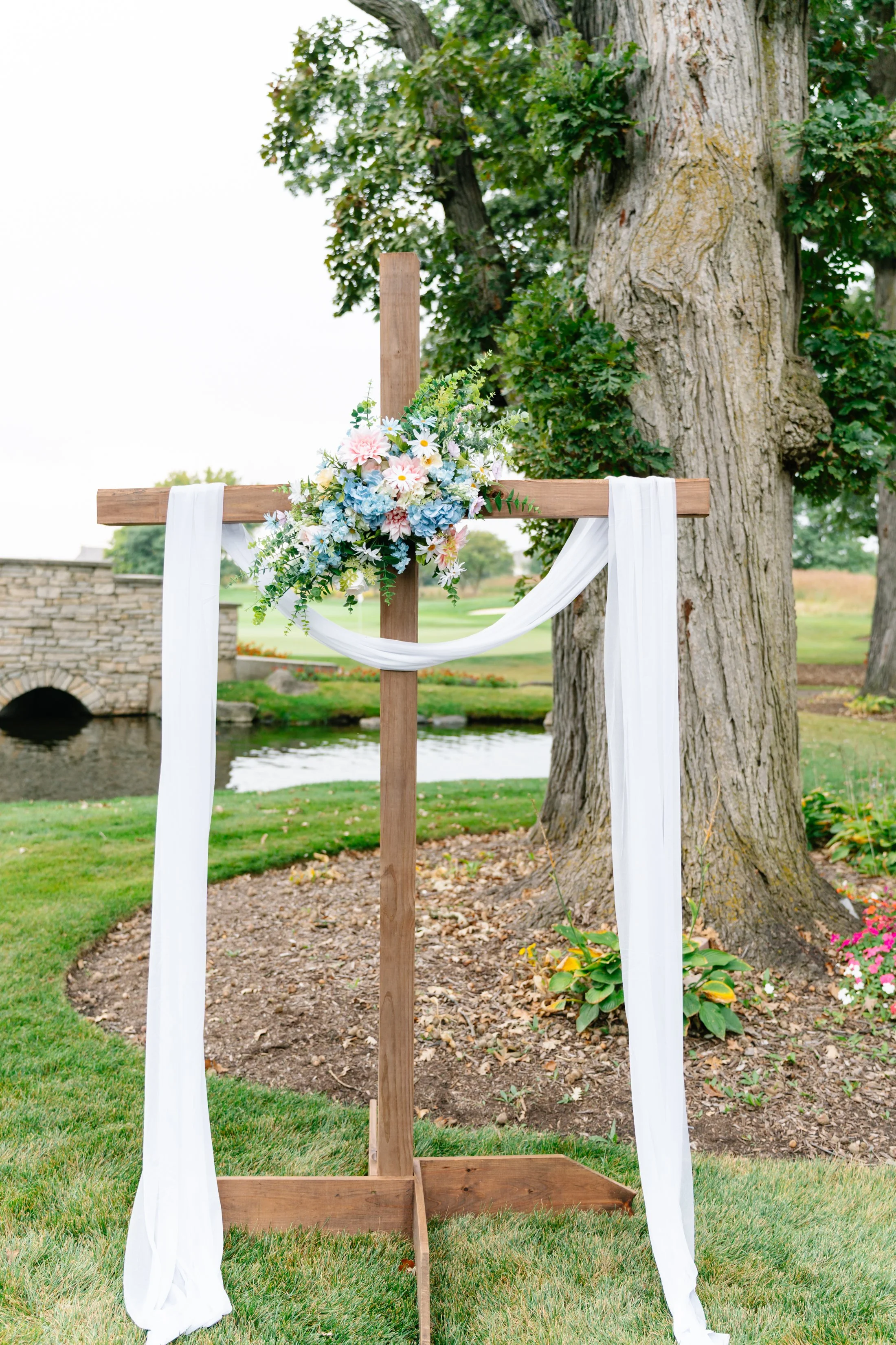 Wooden wedding arch decorated with white fabric and a floral arrangement of pink, white, and blue flowers, set outdoors near a large tree with a grassy area and pond in the background.