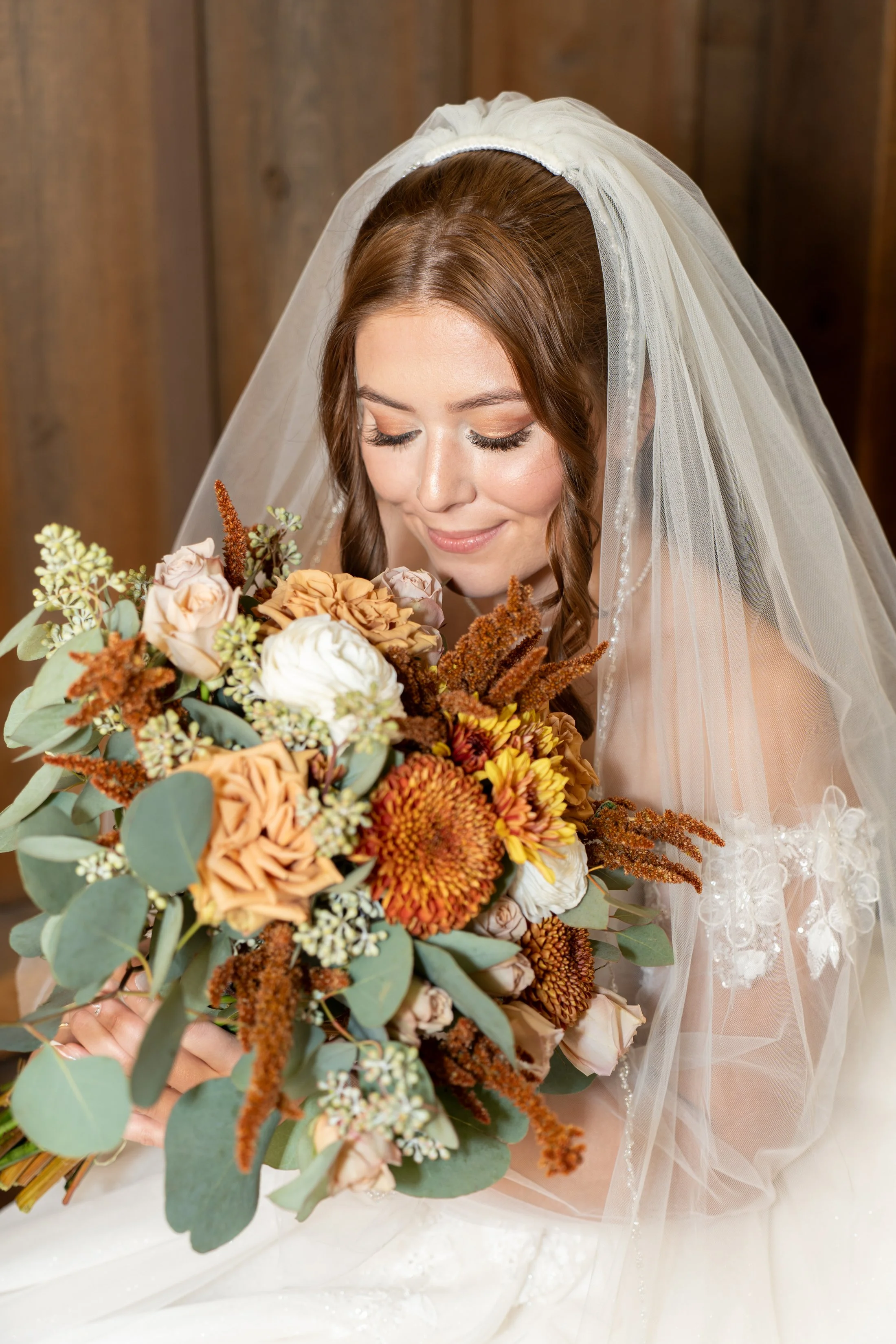 A bride with red hair and a white wedding dress, wearing a veil, holding a large bouquet of mixed flowers including roses, dahlias, and greenery, with a wooden background.