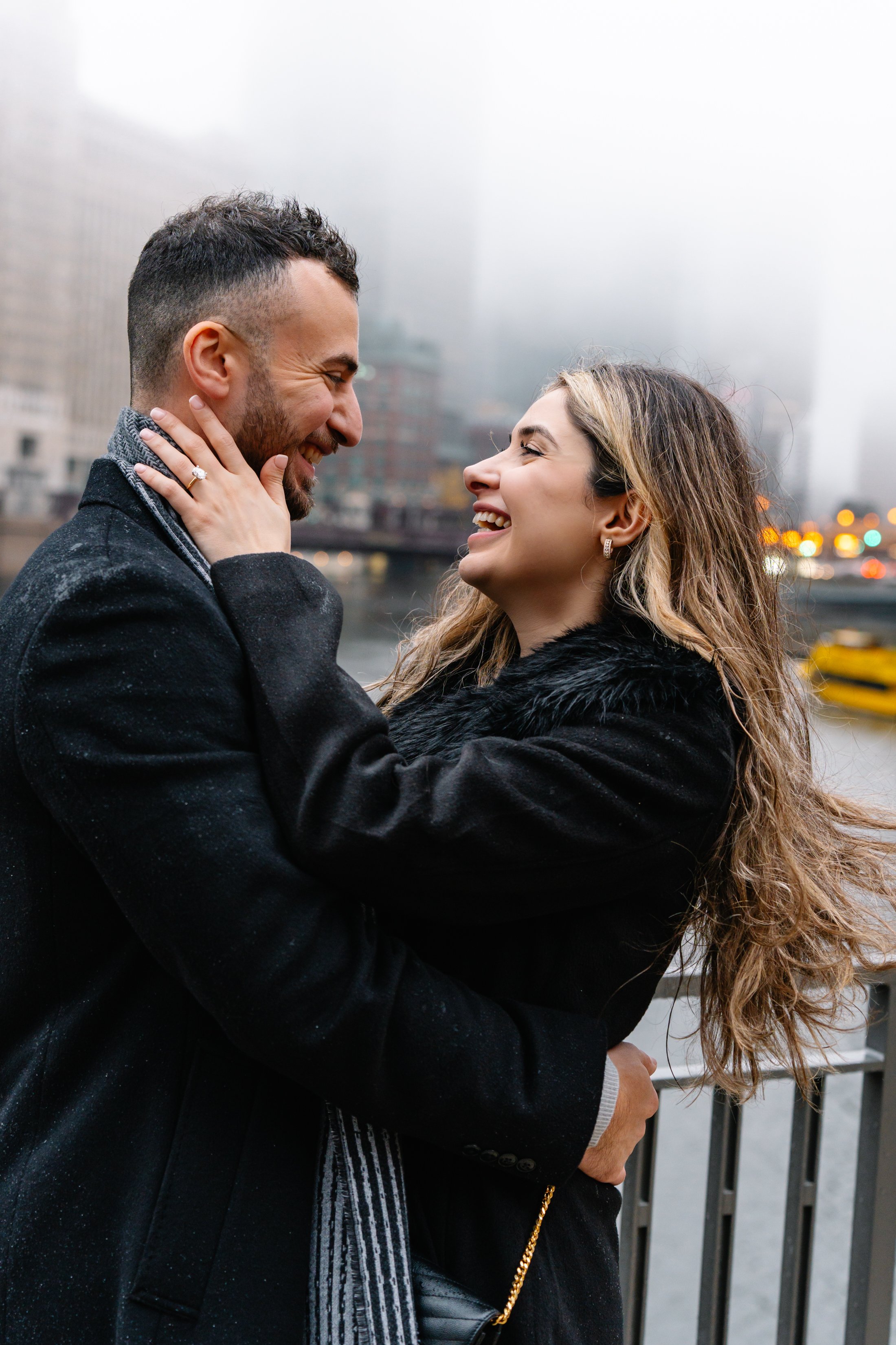 A couple is smiling and embracing outdoors in a city with high-rise buildings and a river in the background on a rainy day.