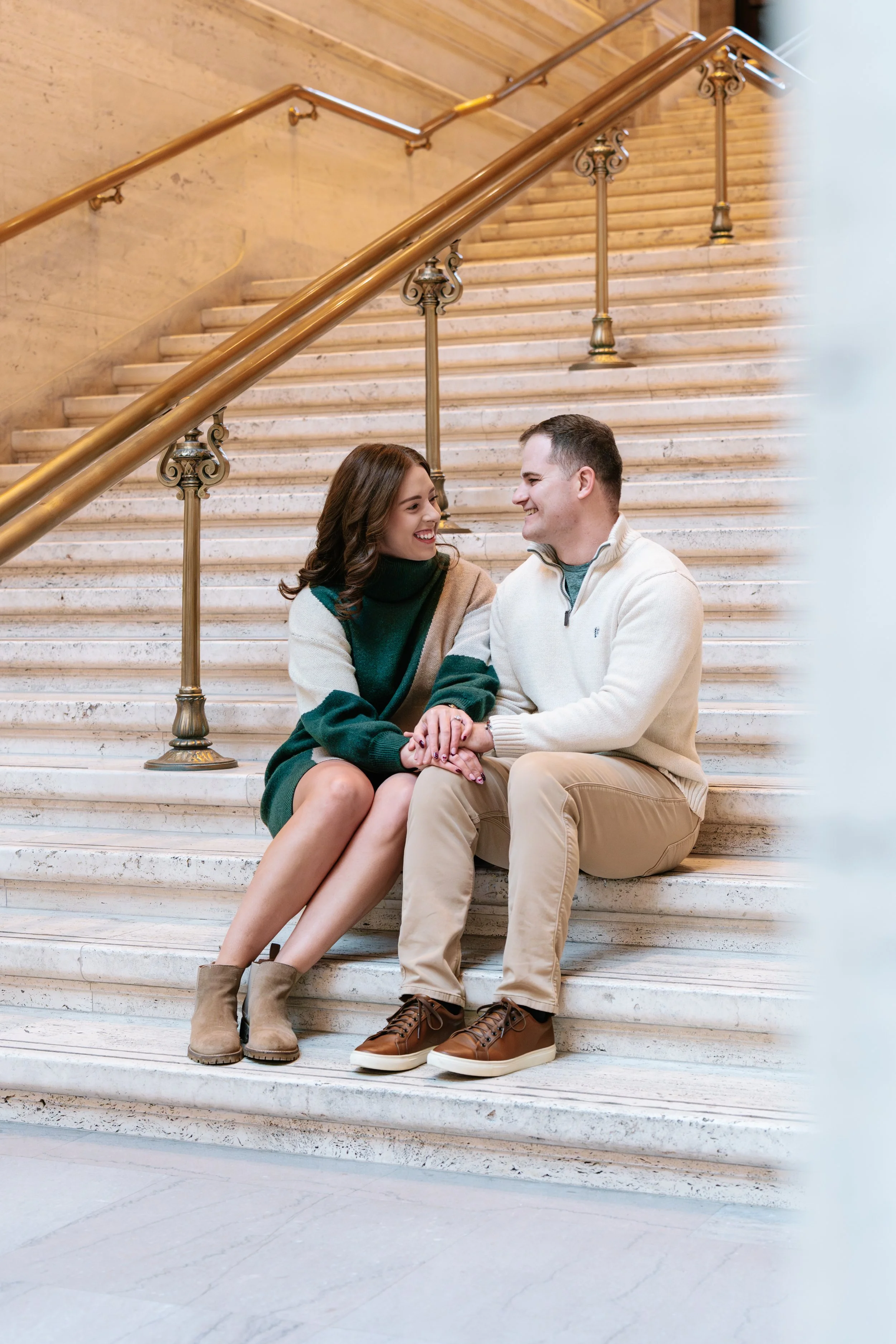 A young couple sitting on marble stairs, holding hands, smiling at each other inside a building with a brass handrail.