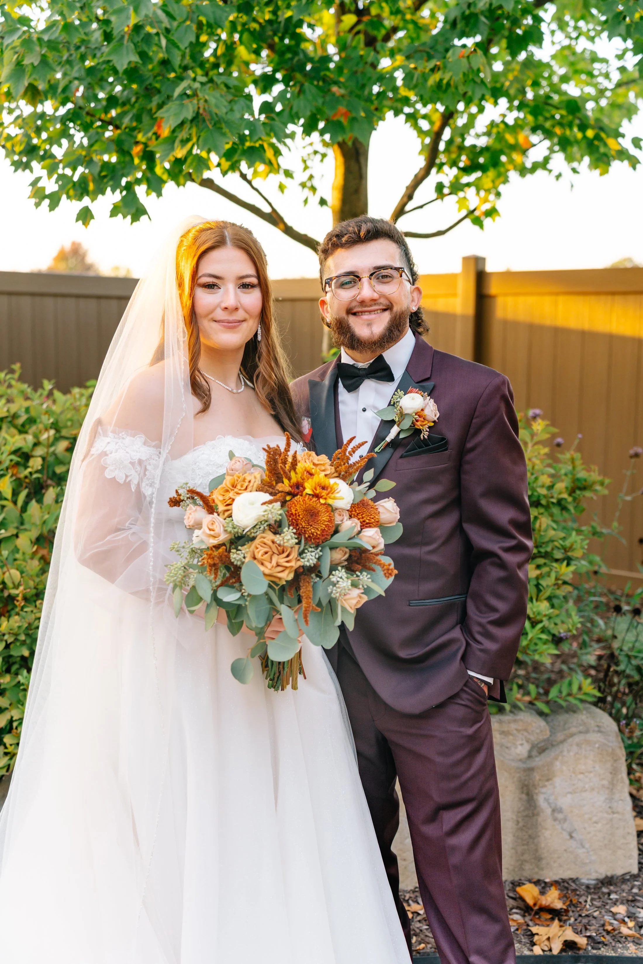 A bride in a white wedding dress and a groom in a dark suit with a bow tie, standing outdoors in front of a tree and a fence during sunset, smiling with the bride holding a large bouquet of orange, peach, and white flowers.