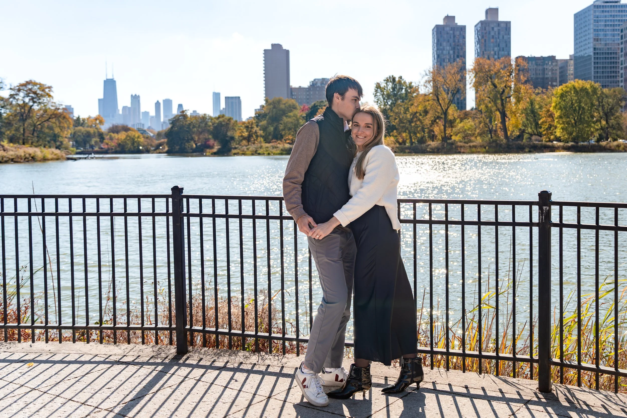 A couple standing by a lake, holding hands and smiling, with city skyscrapers in the background on a sunny day.