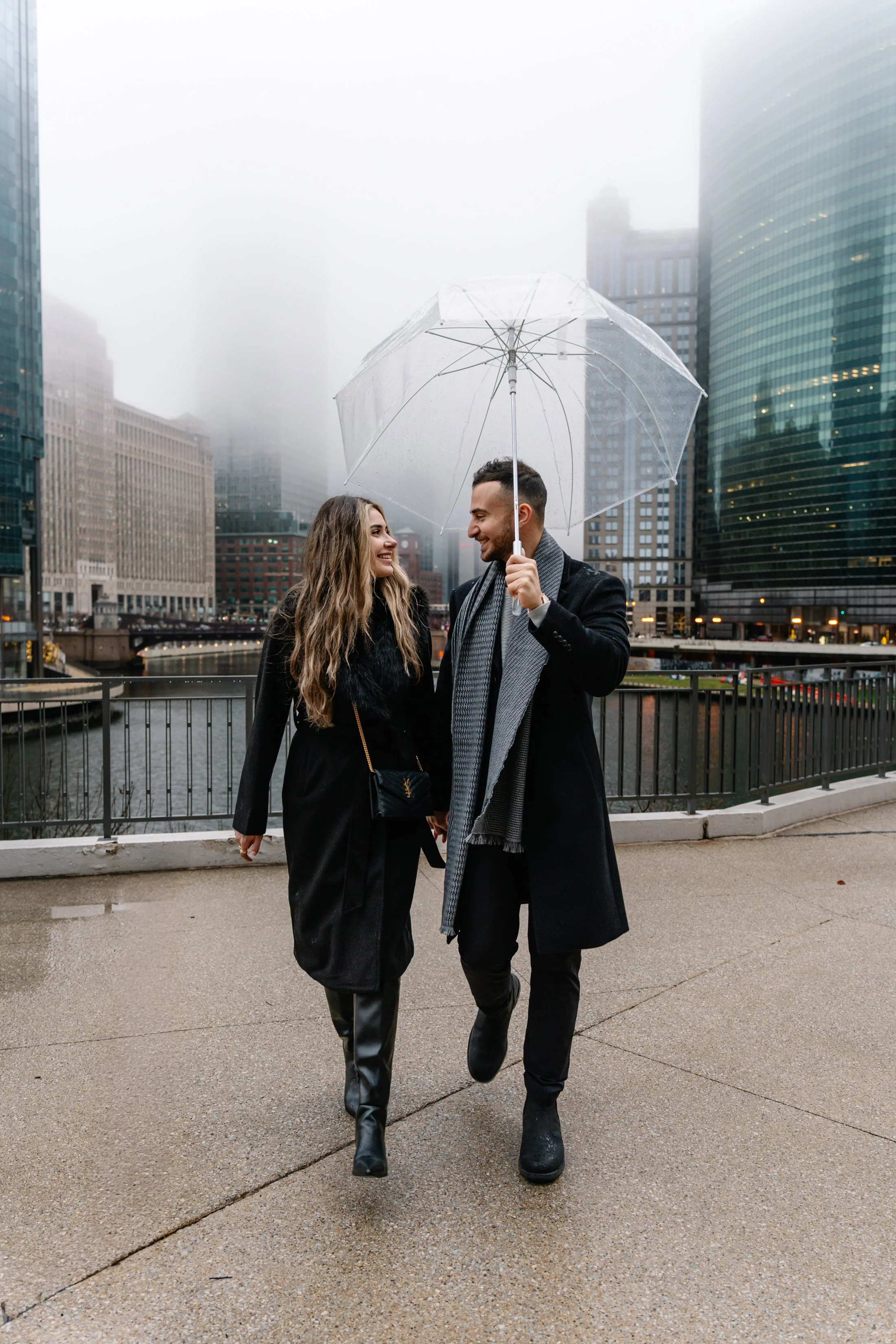 A couple walking in the city on a rainy day, sharing an umbrella, with tall office buildings and a river in the background.