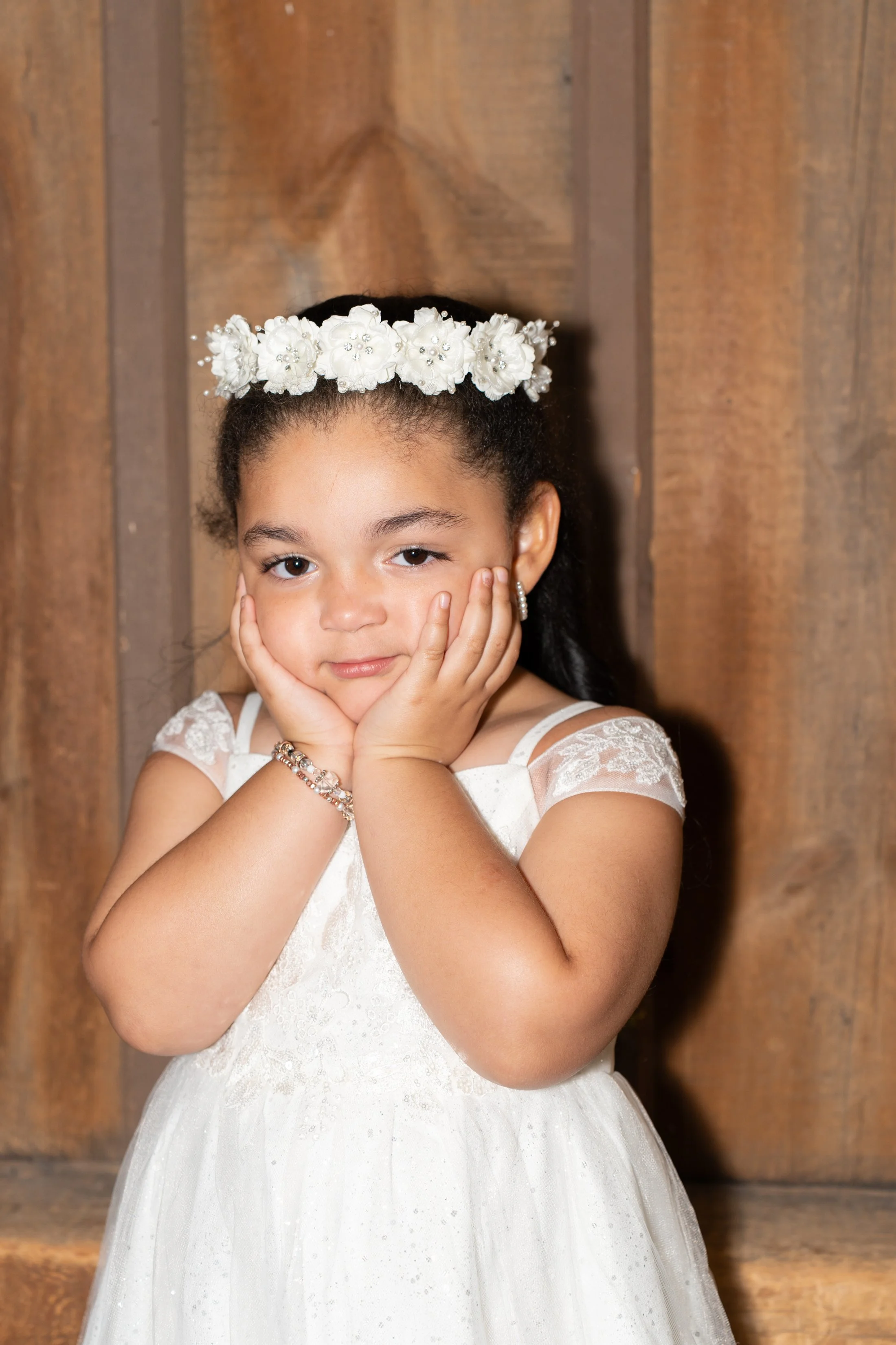 Young girl wearing a white dress with lace details, a white floral headband, and jewelry, standing against a wooden background with her hands resting on her cheeks.