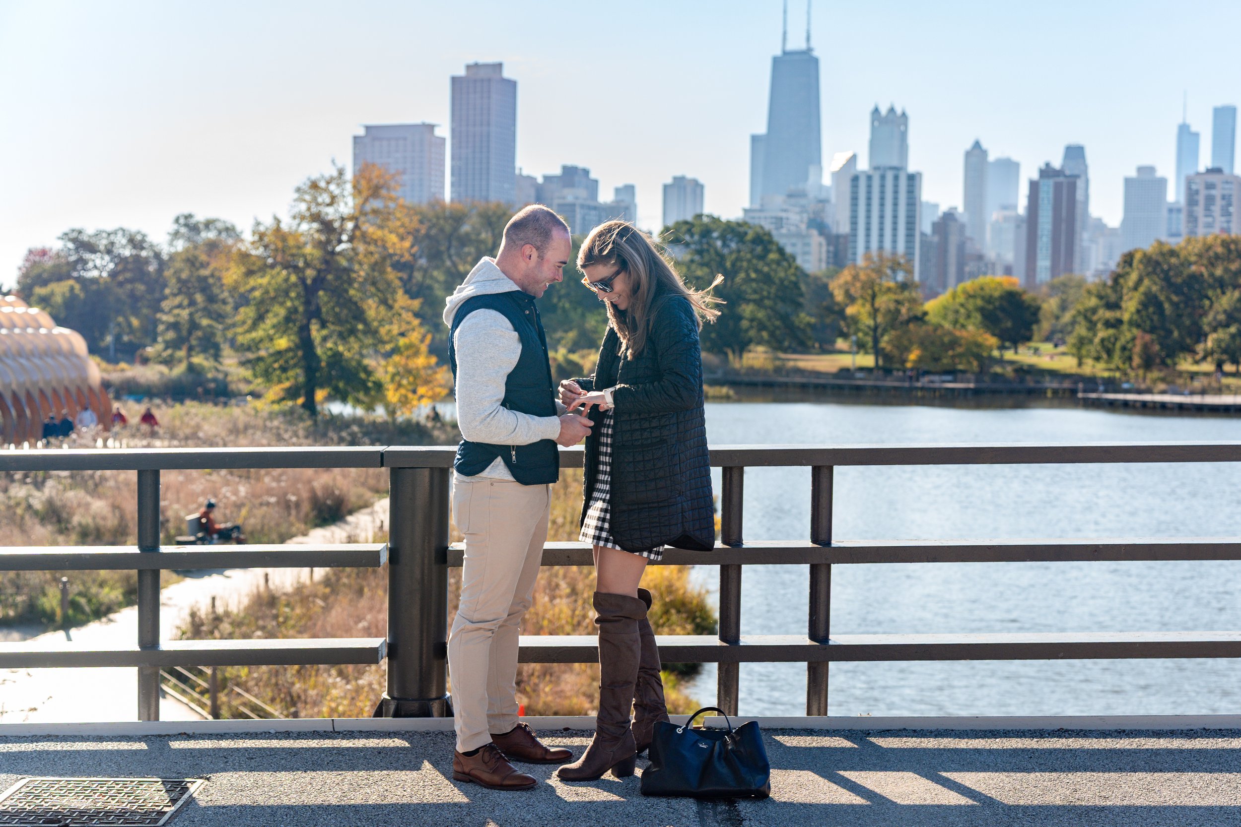 A couple standing on a bridge, looking at a phone together with a city skyline and water in the background.