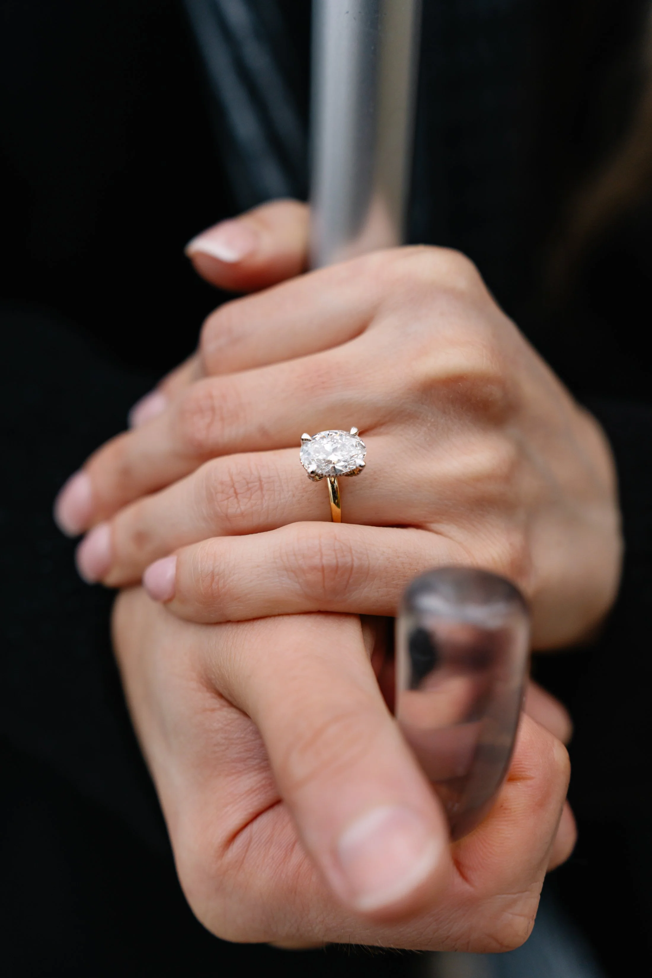 Close-up of a woman's hand with a large diamond ring on her ring finger, holding onto a metallic object, against a dark background.