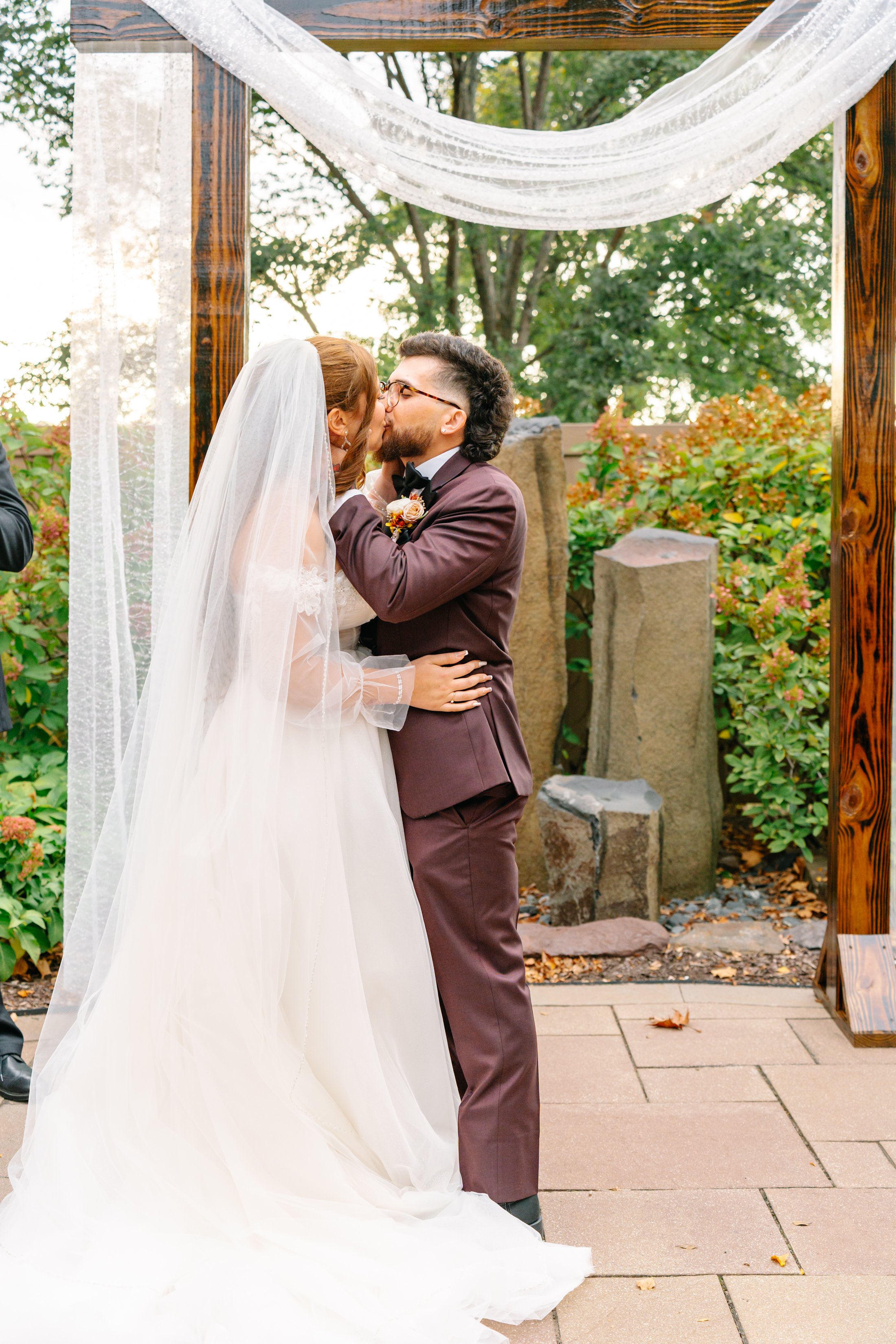 A couple kissing at their outdoor wedding ceremony, with the bride in a white gown and veil and the groom in a dark suit, under a wooden arch decorated with white fabric. The background features green foliage and stones.