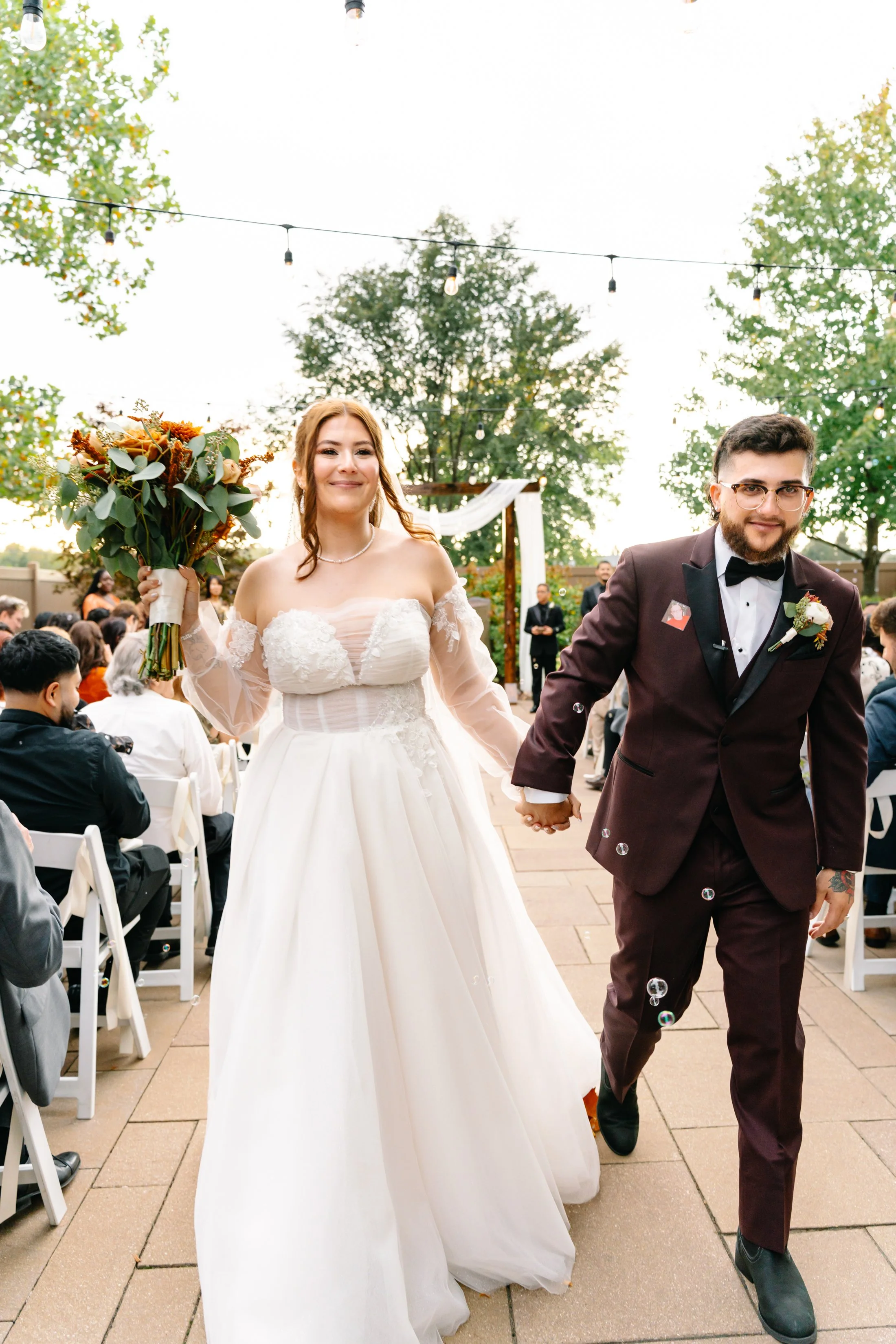 A bride and groom walking hand in hand after their wedding ceremony outdoors, surrounded by wedding guests, with trees and string lights overhead.