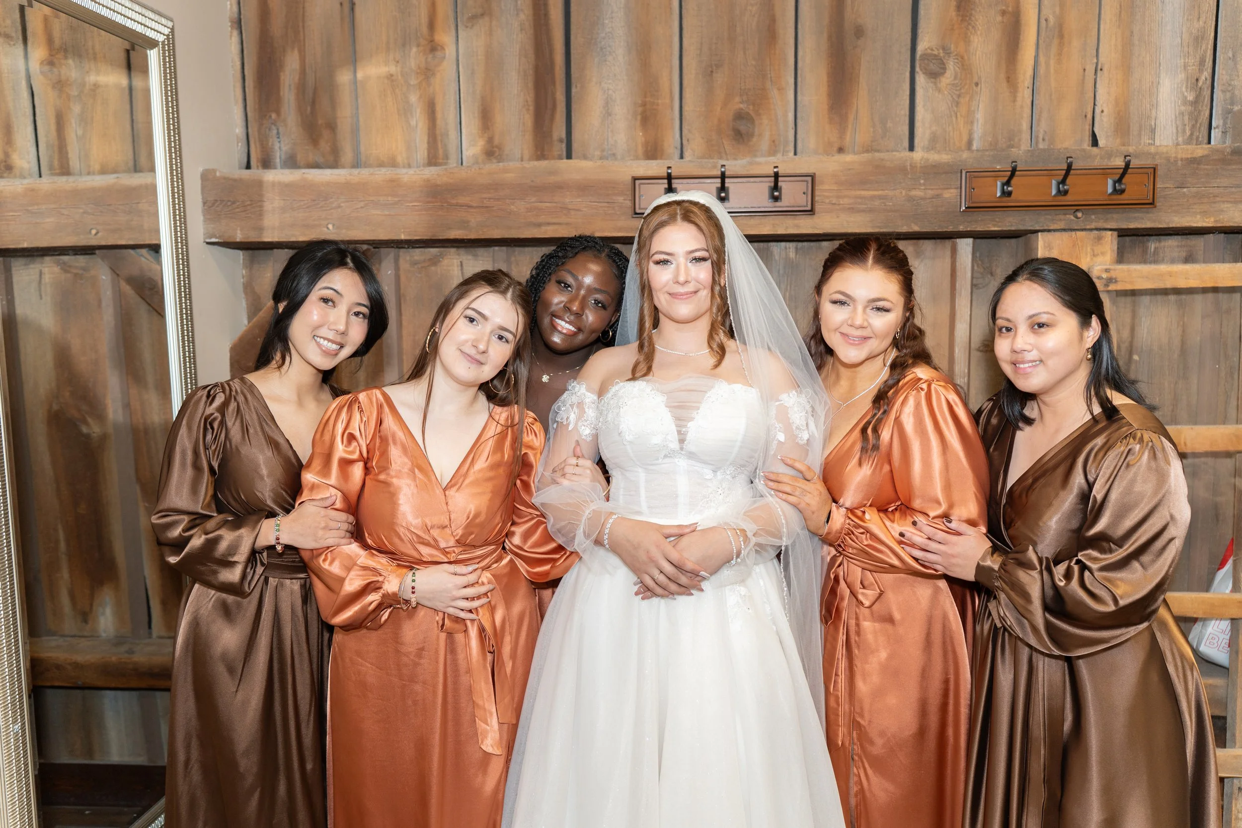 Bride in a white wedding dress with veil, surrounded by five women in satin robes, standing in front of a wooden wall.